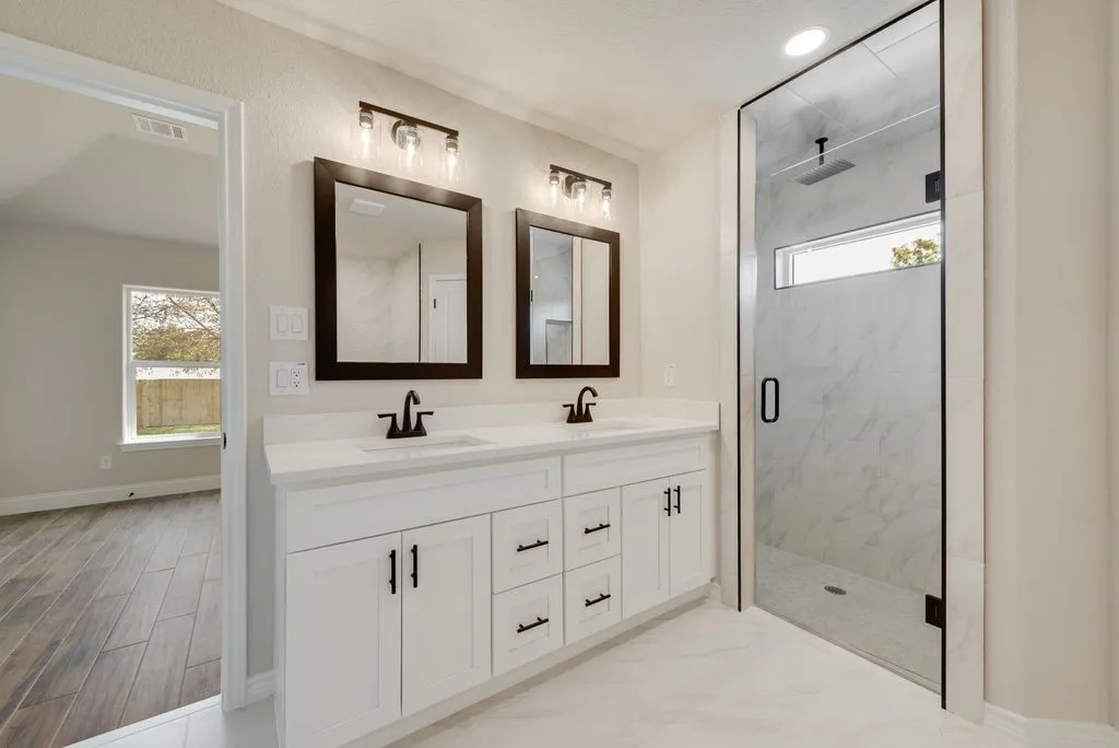 Dual-sink vanity with white cabinetry and light-toned countertop, framed vanity mirrors, dual three-light fixtures, and a walk-in shower with stone-surround tile and a glass door