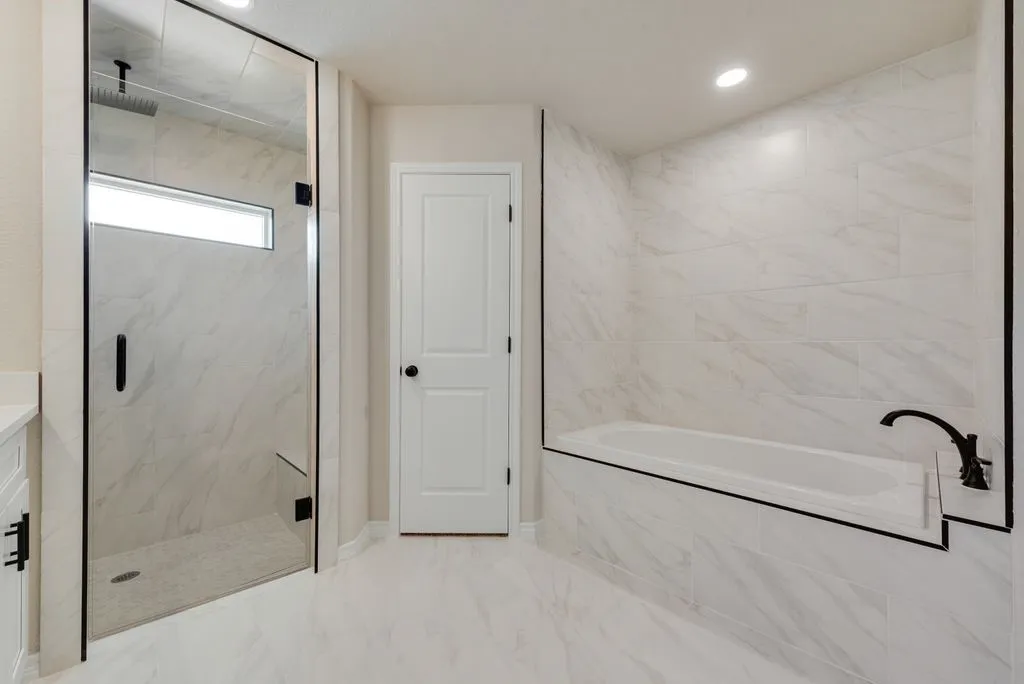 Walk-in glass-enclosed shower featuring stone-finish wall tile, built-in bench seating, and matte black fixtures, accompanied by a soaking tub with matching surround and a dedicated shower head