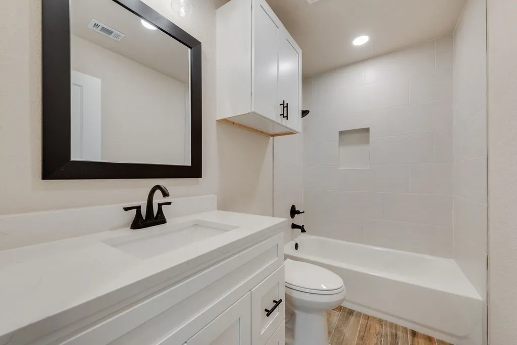 White vanity with integrated countertop, matte black fixtures, framed wall mirror, white cabinetry, and tiled shower surround with recessed niche