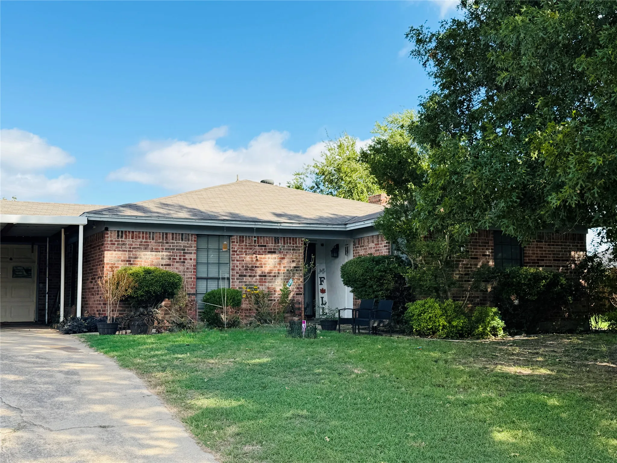 Single-story brick exterior featuring a hip roof, concrete driveway, attached carport, and mature trees