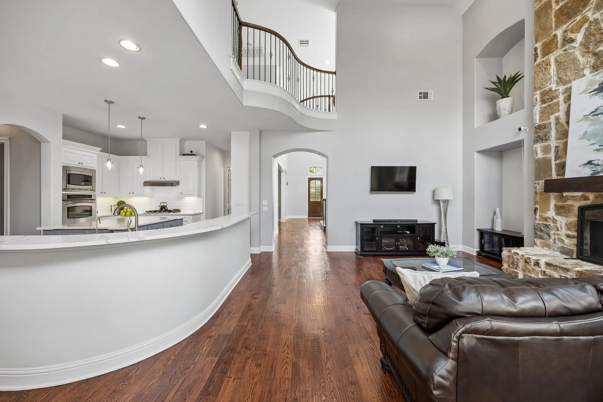 Light-filled living area with soaring ceilings, rich wood floors, and seamless flow to the kitchen, anchored by a striking stone fireplace.