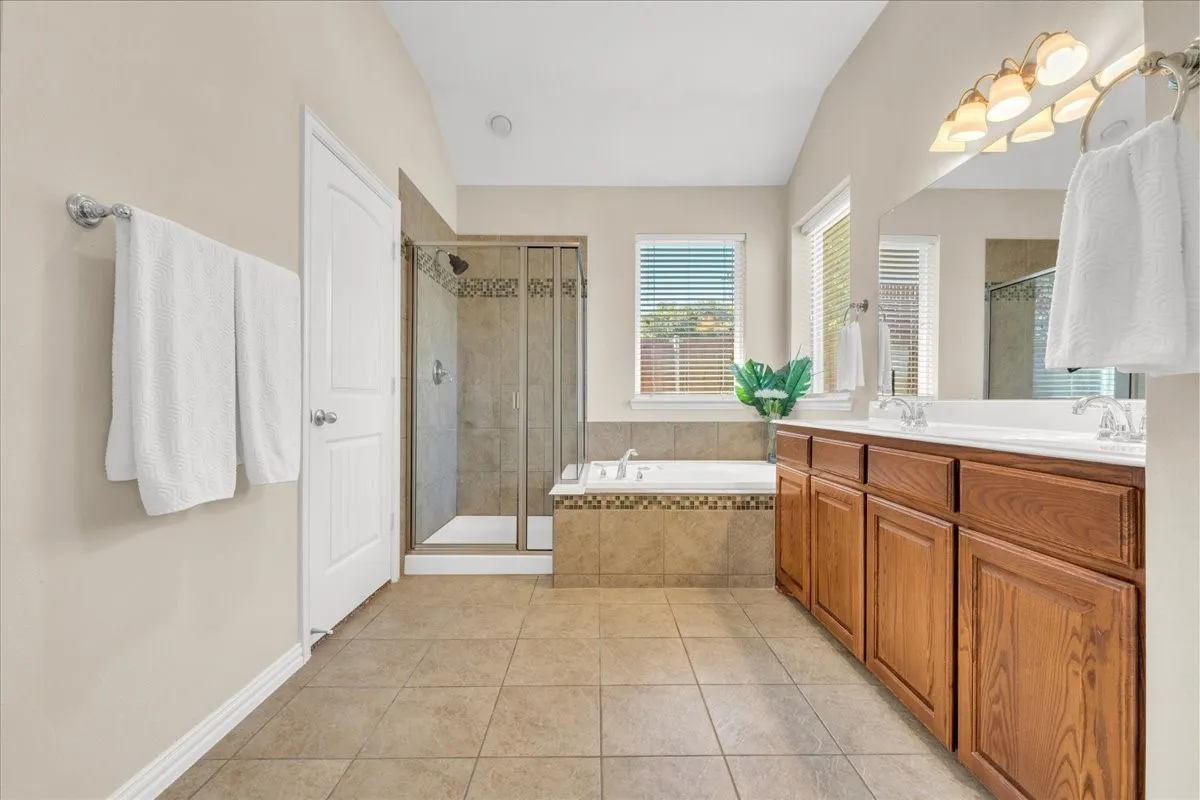 Spacious bathroom featuring a dual vanity with wood-finish cabinetry, a tiled soaking tub, a separate glass-enclosed shower, and tile flooring