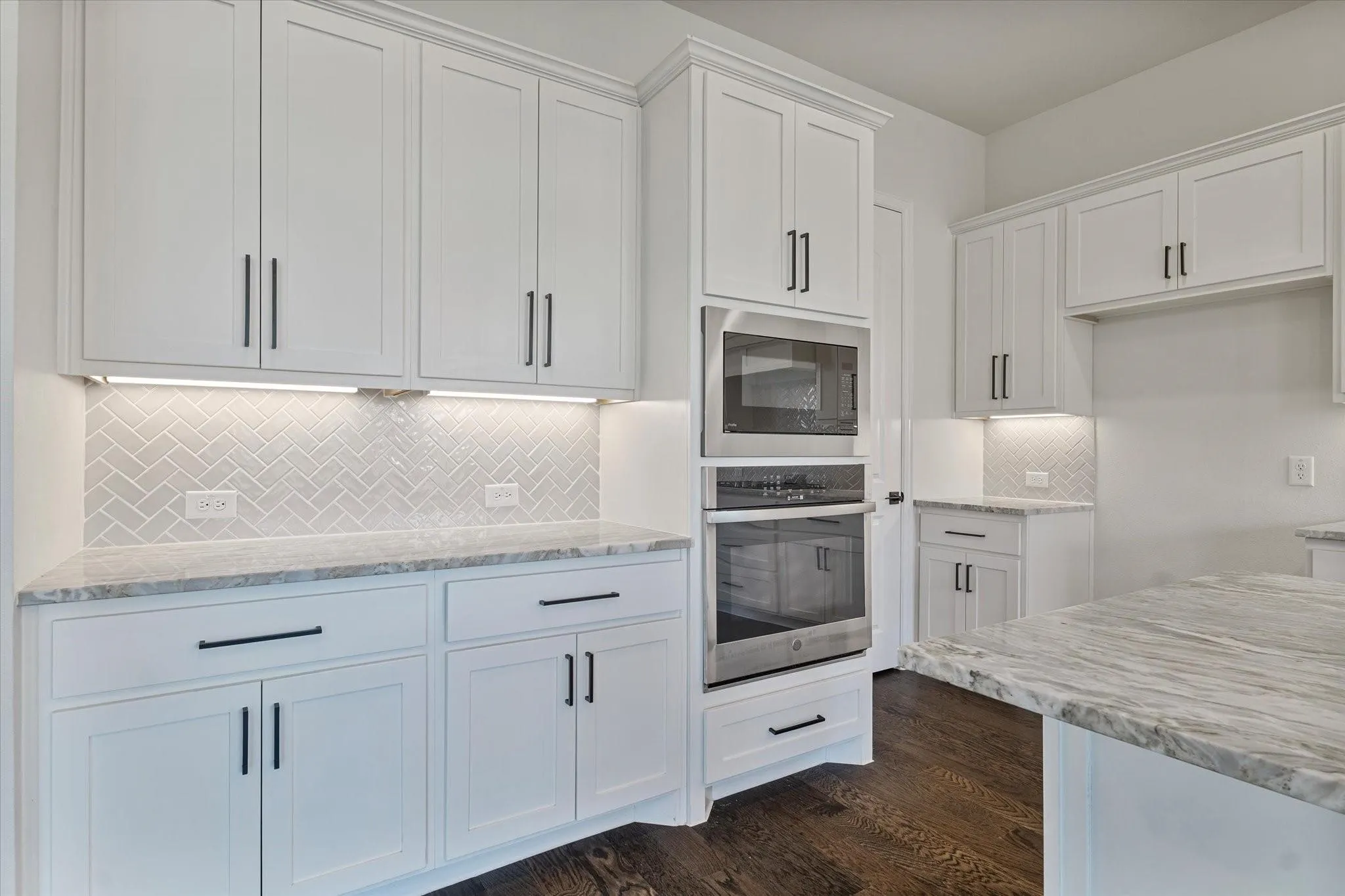 Kitchen featuring white Shaker-style cabinetry, herringbone-patterned tile backsplash, light-toned stone countertops, built-in stainless steel double ovens, and dark wood-finish flooring