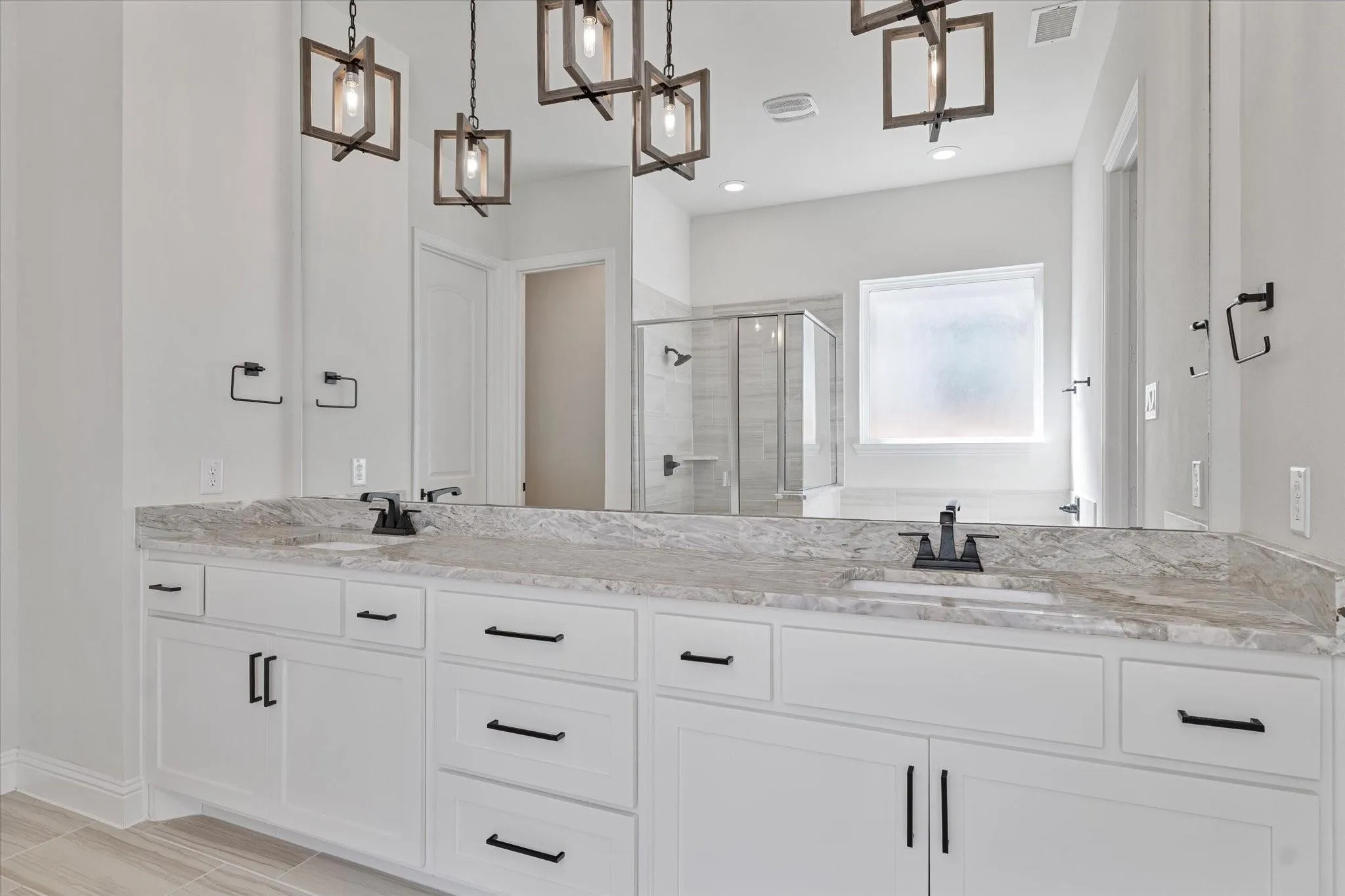 Dual-sink vanity with stone-finish countertop, white Shaker-style cabinetry, matte black hardware, geometric pendant lighting, and a walk-in glass-enclosed shower