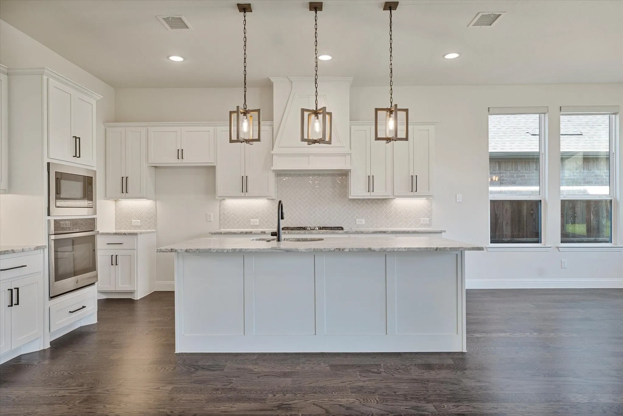 Kitchen featuring white cabinetry, center island with stone countertop, herringbone tile backsplash, stainless steel double wall ovens, and three geometric pendant lights over the island