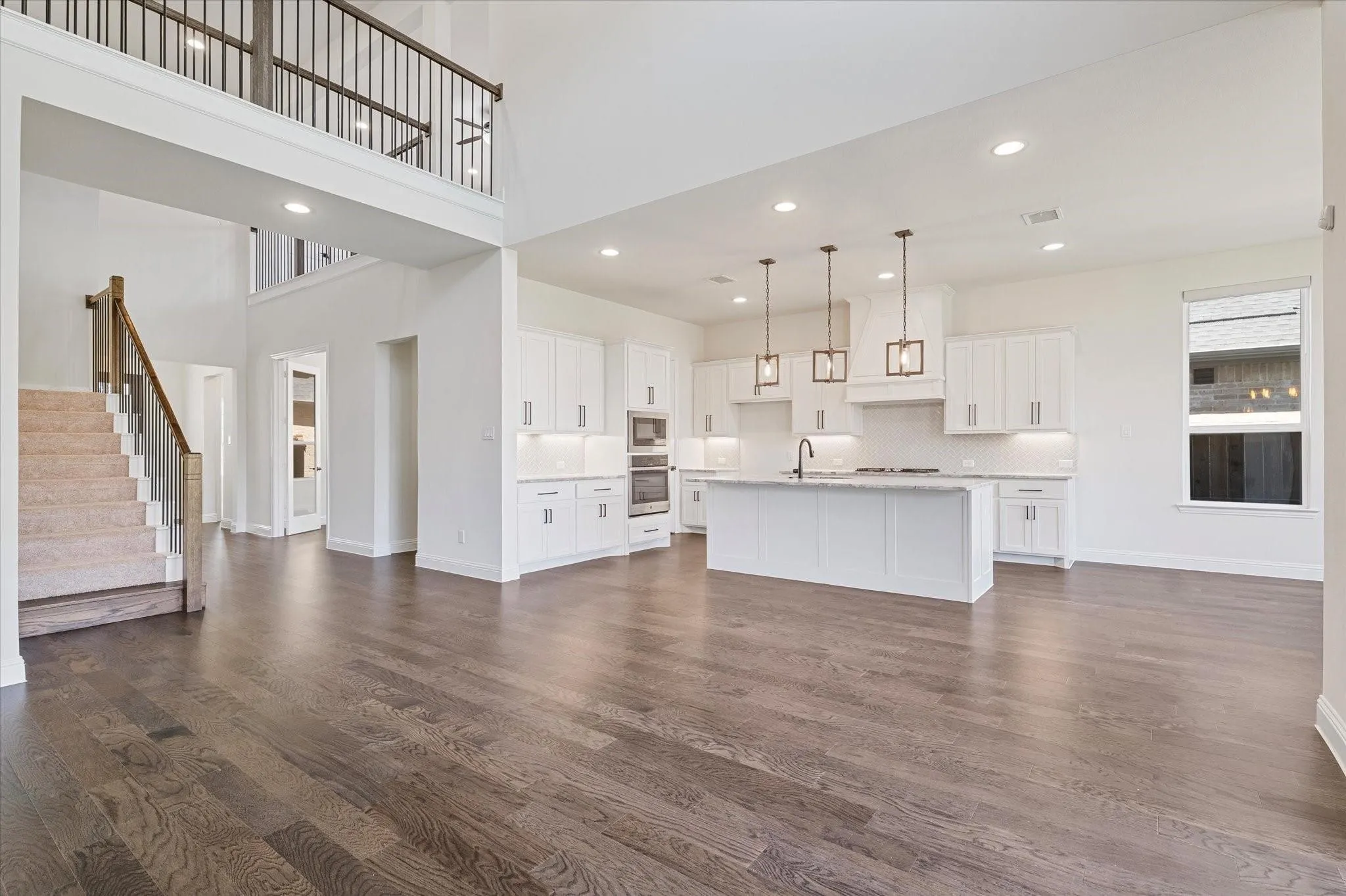 Open-concept interior featuring wood-finish flooring, a double-height ceiling, a center island with pendant lighting, white shaker-style cabinetry, and a carpeted staircase with a wood railing