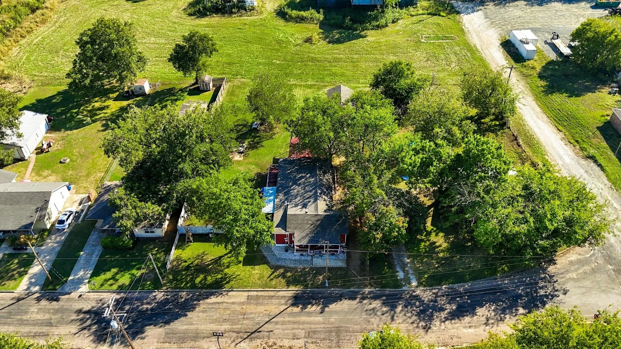 Aerial perspective of property featuring an expansive grass lot, mature trees, a detached storage building, and a paved street