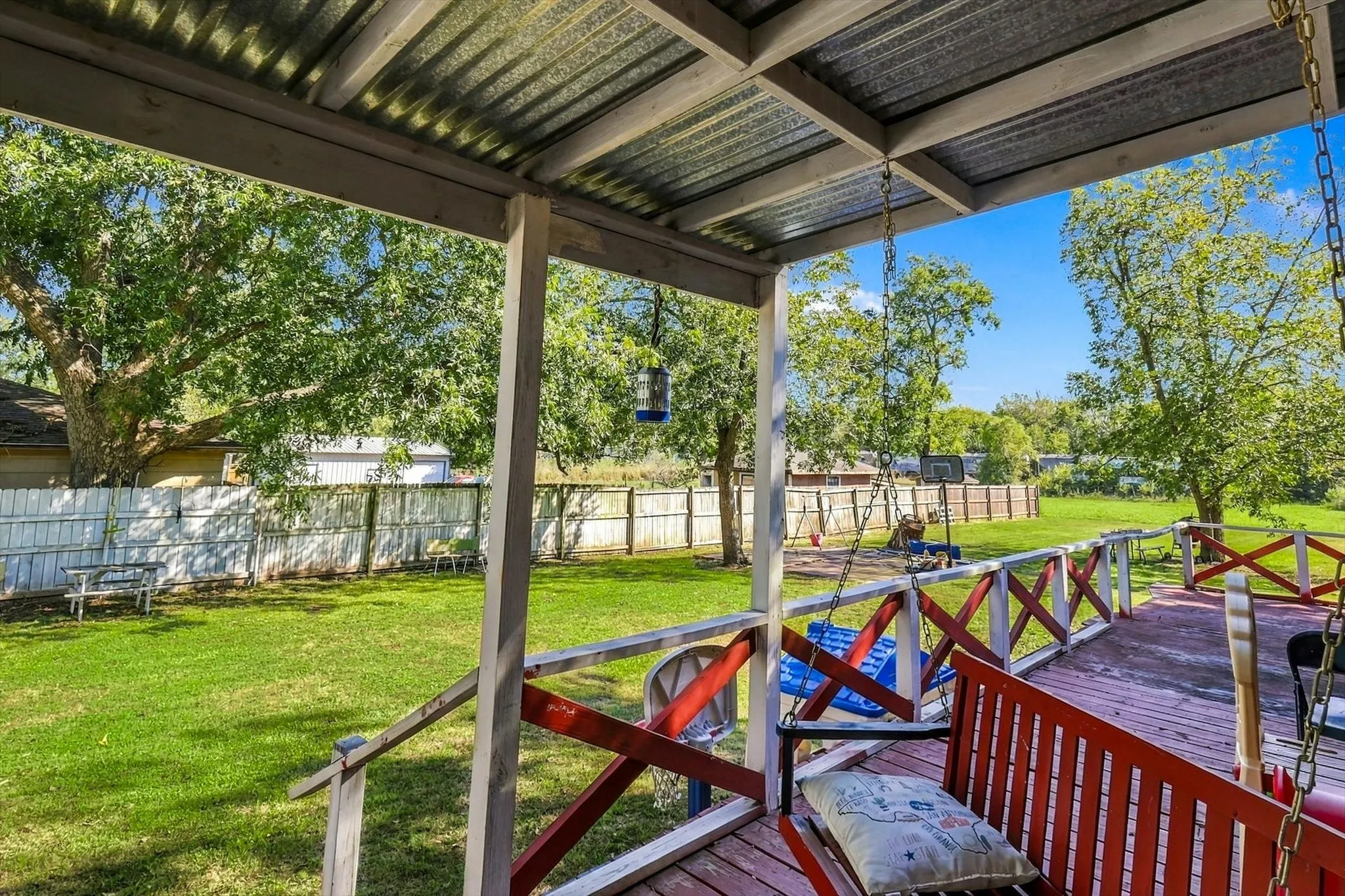 Covered deck featuring a corrugated metal roof, wood-finish decking, and painted cross-buck railing, overlooking a fenced backyard with mature trees