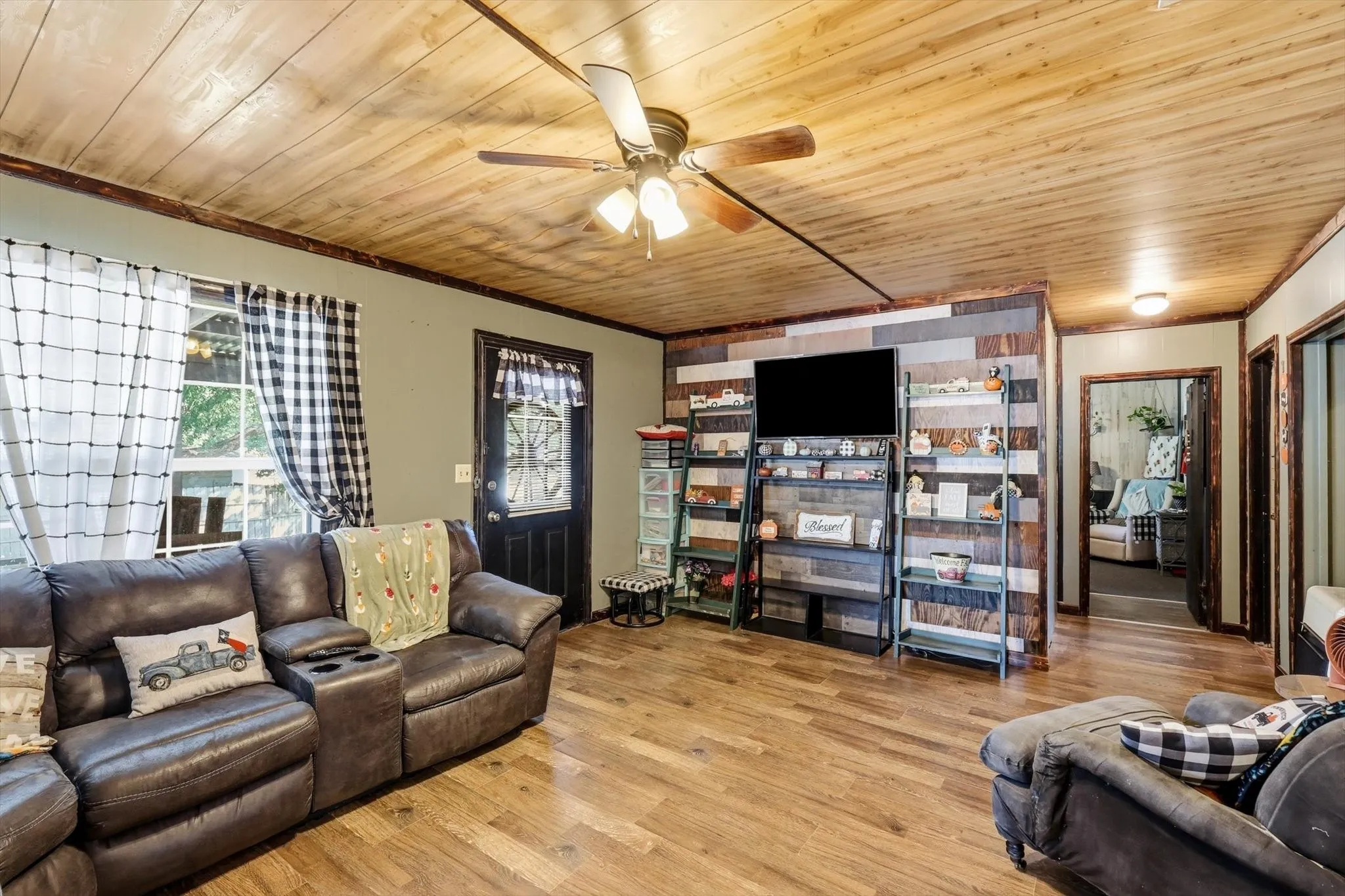 Living area featuring wood-plank ceiling, wood-finish flooring, multi-tonal wood accent wall, and ceiling fan with integrated lighting