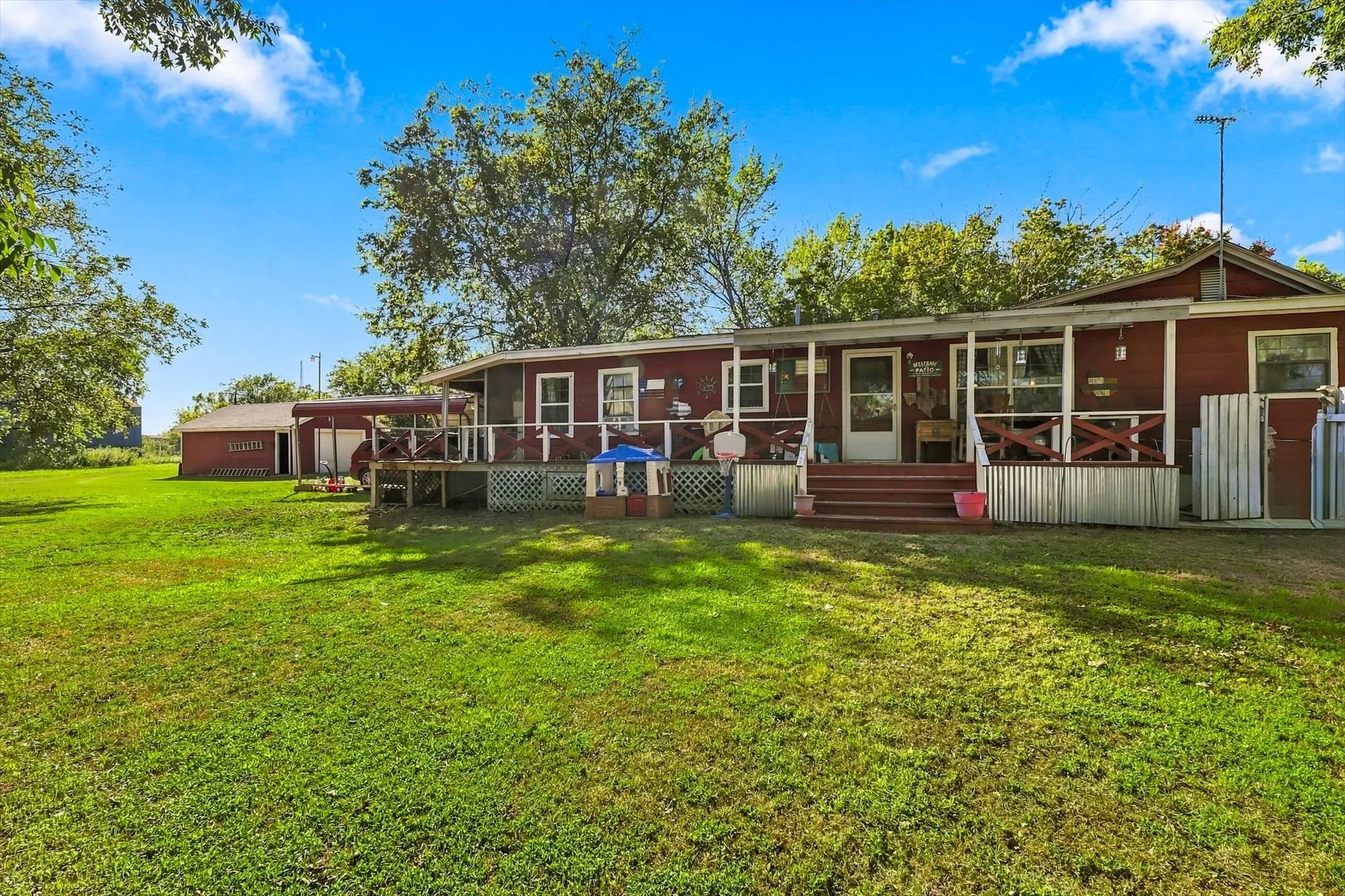 Red exterior residence featuring a covered wooden porch with cross-buck railings, corrugated metal skirting, and an expansive lawn with a detached metal-sided storage structure