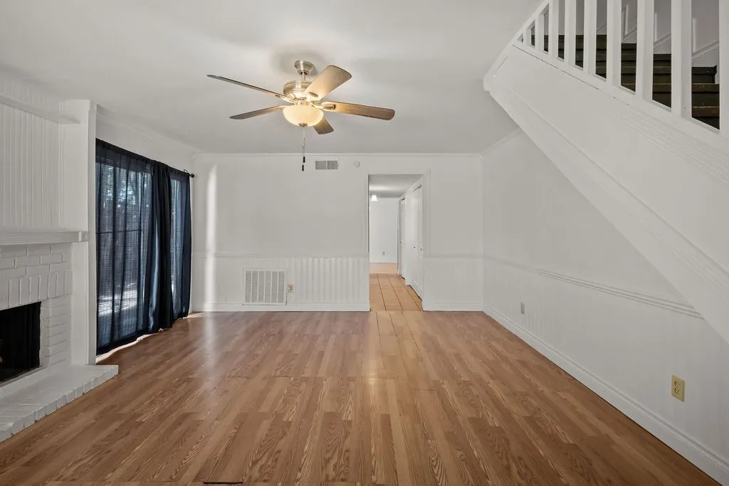 Living area featuring wood-finish flooring, a painted brick fireplace, beadboard wainscoting, a ceiling fan with integrated lighting, and a staircase with white balusters