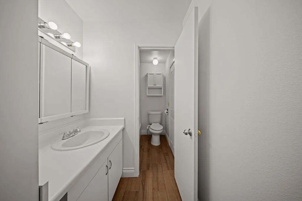 Bathroom featuring a white vanity cabinet with an integrated sink, a mirrored medicine cabinet, a multi-bulb light fixture, wood-finish flooring, and a wall-mounted storage cabinet above the toilet
