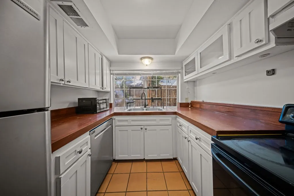 Kitchen featuring white shaker-style cabinetry with glass-front uppers, wood-finish countertops, stainless steel appliances, a double-basin sink with a matte black pull-down faucet, and ceramic tile flooring