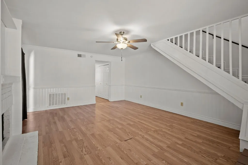 Interior space featuring wood-finish flooring, wainscoting, a fireplace with a painted brick surround, a ceiling fan with integrated lighting, and a staircase with white balusters