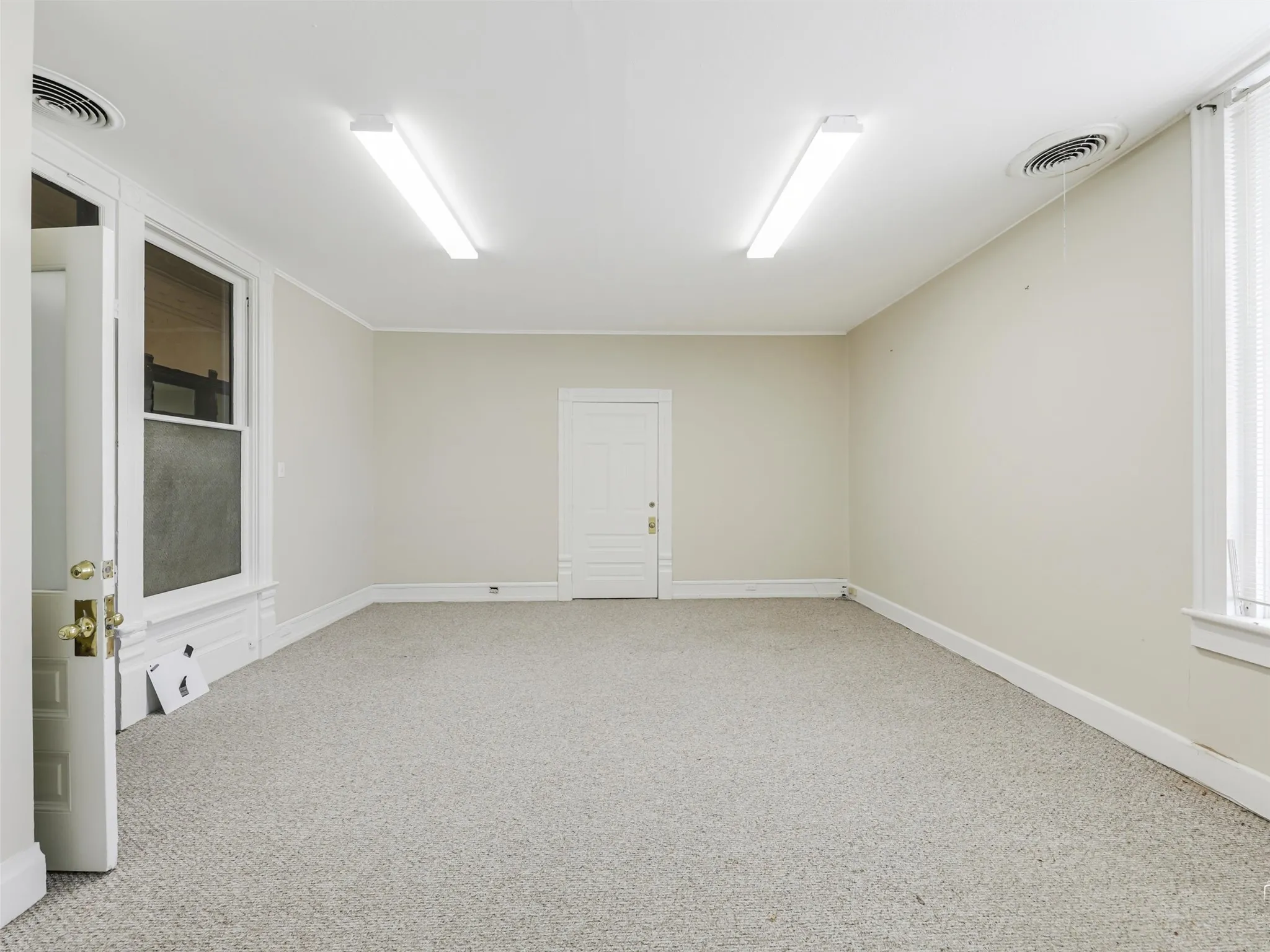 Interior room featuring neutral wall paint, carpeted flooring, two linear overhead light fixtures, and white paneled interior doors with brass-finish hardware