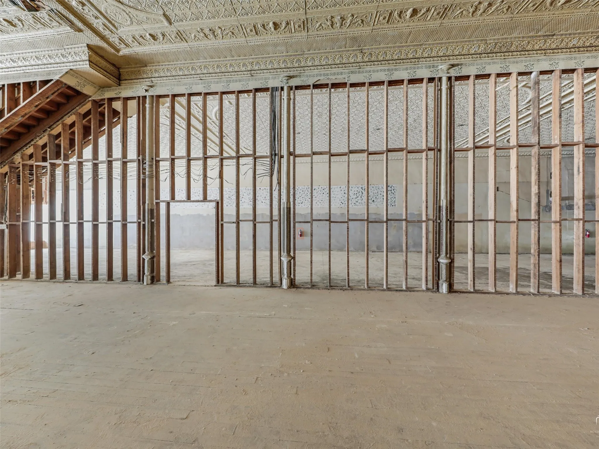 Open-plan interior featuring ornate pressed-tin ceilings, exposed wood wall framing, decorative metal columns, and an unfinished staircase
