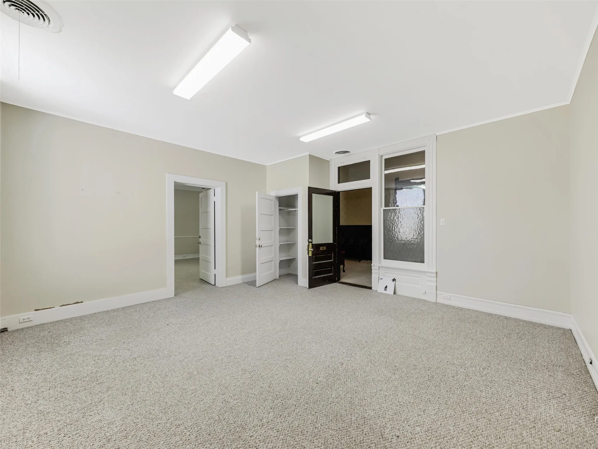 Spacious room featuring neutral carpeting, multi-panel doors, built-in shelving, linear overhead lighting, and a window with a fixed transom