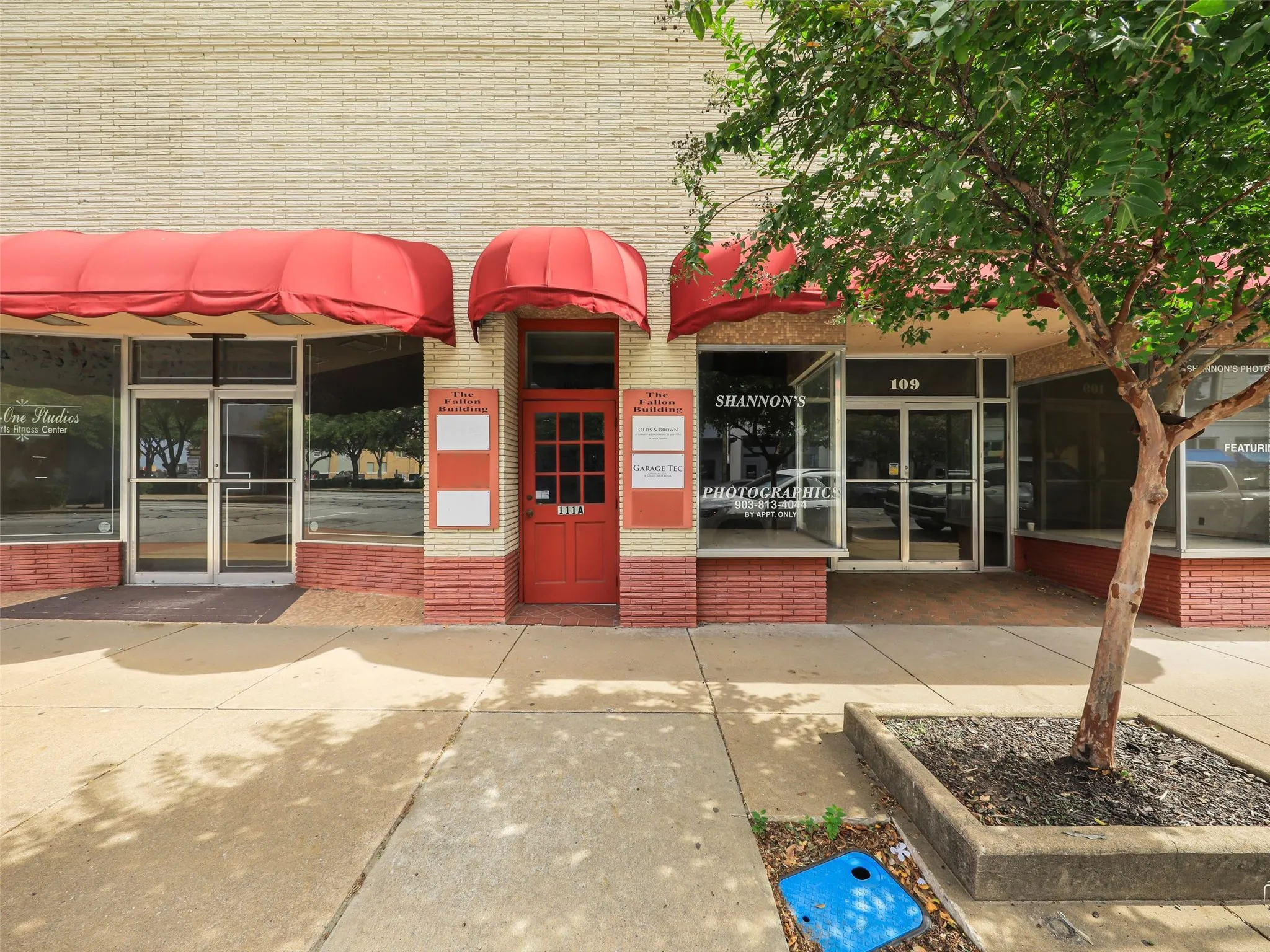 Commercial building exterior featuring light-colored brick facade, red-tinted masonry base, multi-pane entry door, glass storefront windows, and fabric awnings