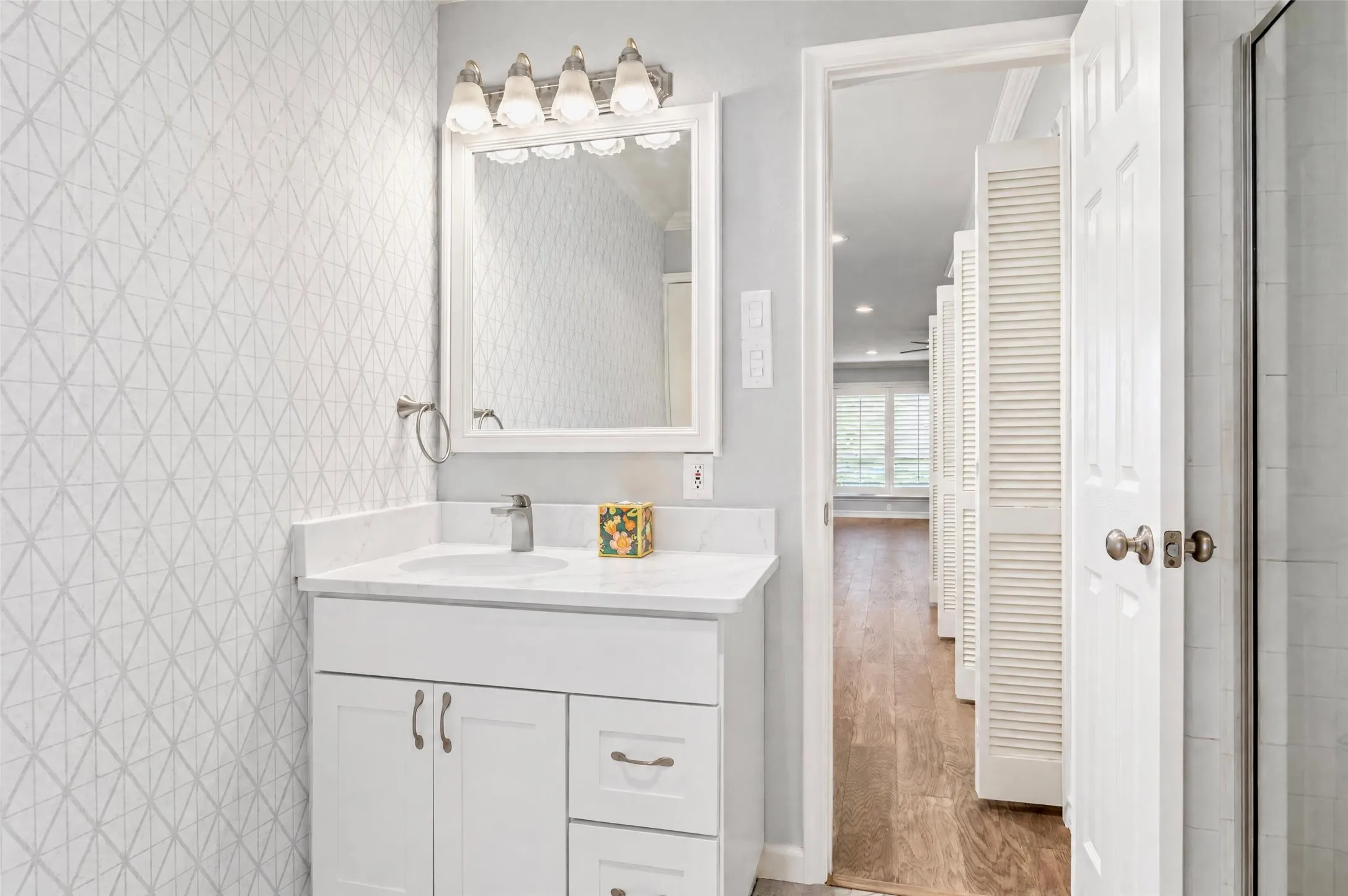 White vanity with stone countertop, undermount sink, geometric patterned wall covering, four-light vanity fixture, and framed wall mirror