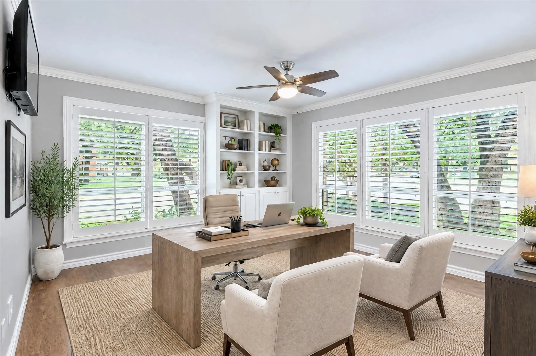 Room featuring wood-finish flooring, built-in floor-to-ceiling shelving, triple-window configuration with white plantation shutters, and a central ceiling fan with integrated lighting