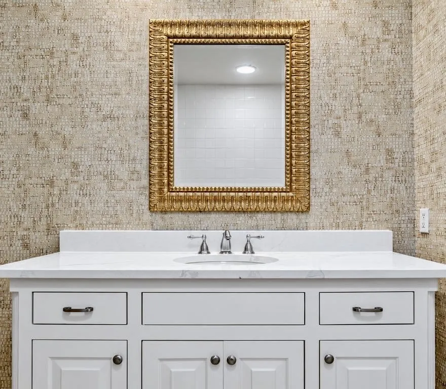 White cabinetry vanity featuring a stone countertop, an integrated oval sink, a three-piece chrome faucet, and textured wall covering, accented by a decorative metallic framed mirror