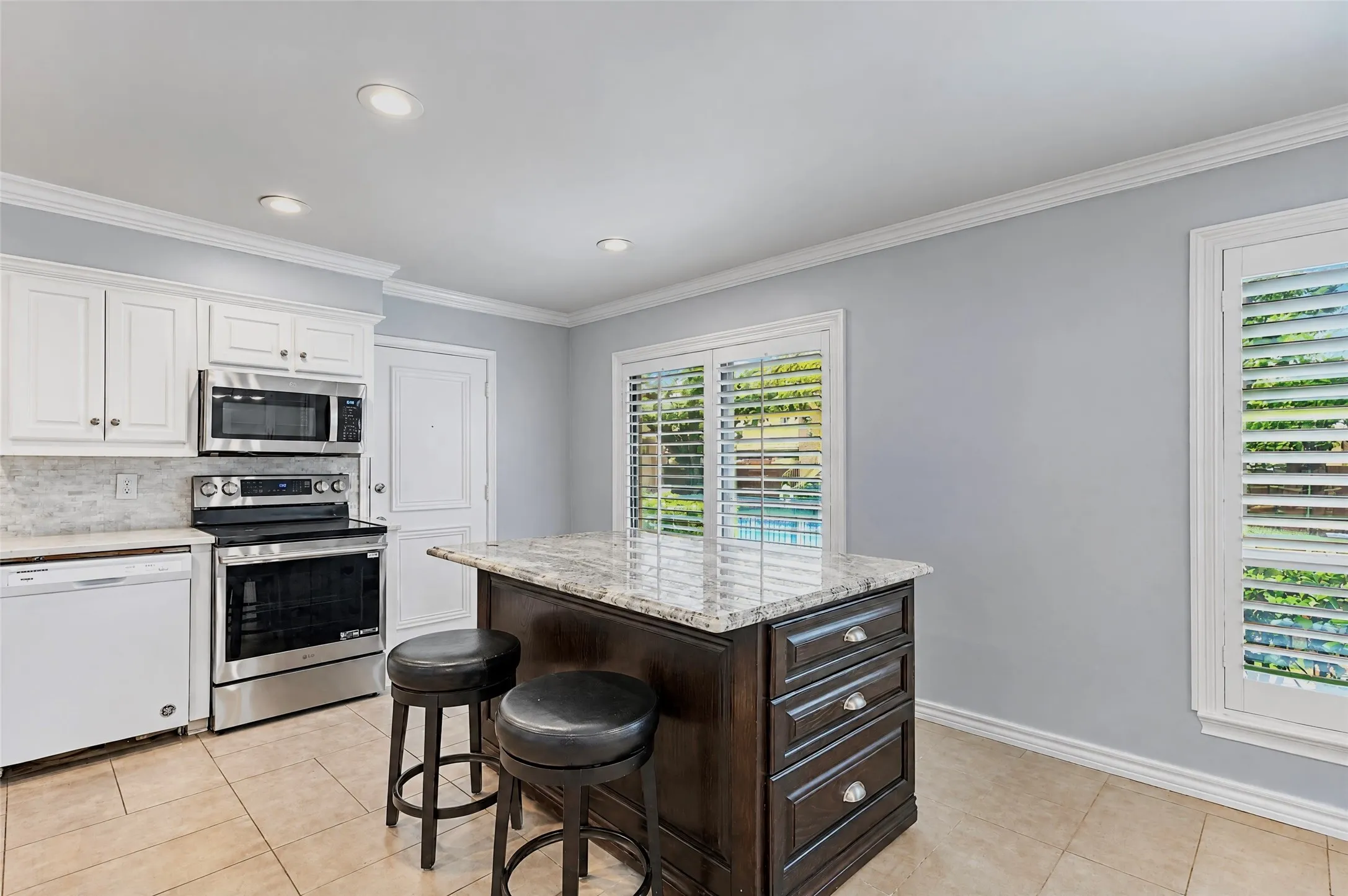 Kitchen featuring a central island with stone countertop, white shaker-style cabinetry, stainless steel appliances, tile flooring, and plantation shutters