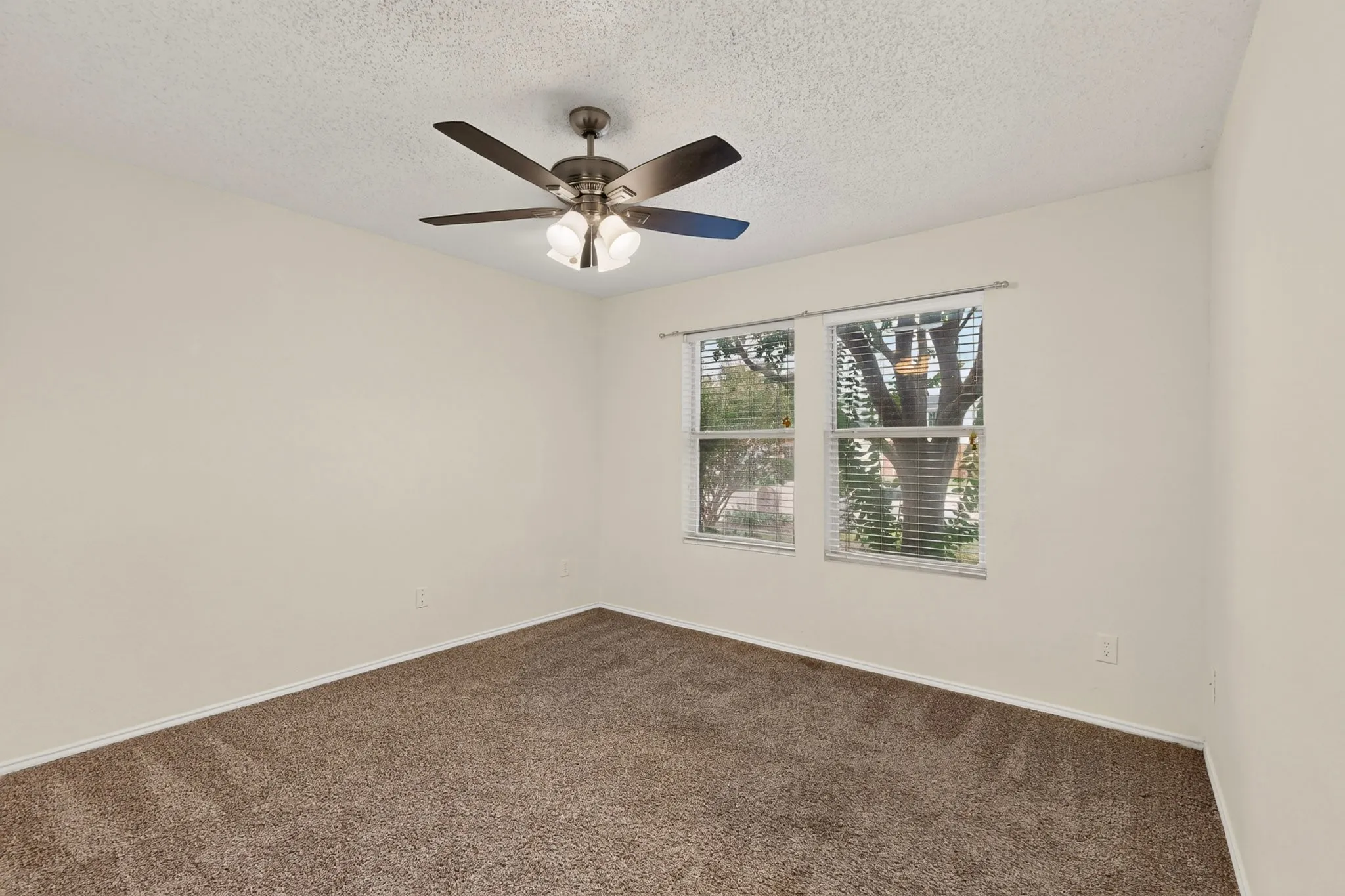 Neutral-toned interior space featuring textured ceiling, carpeting, multi-blade ceiling fan with integrated lighting, and dual windows with metal rods