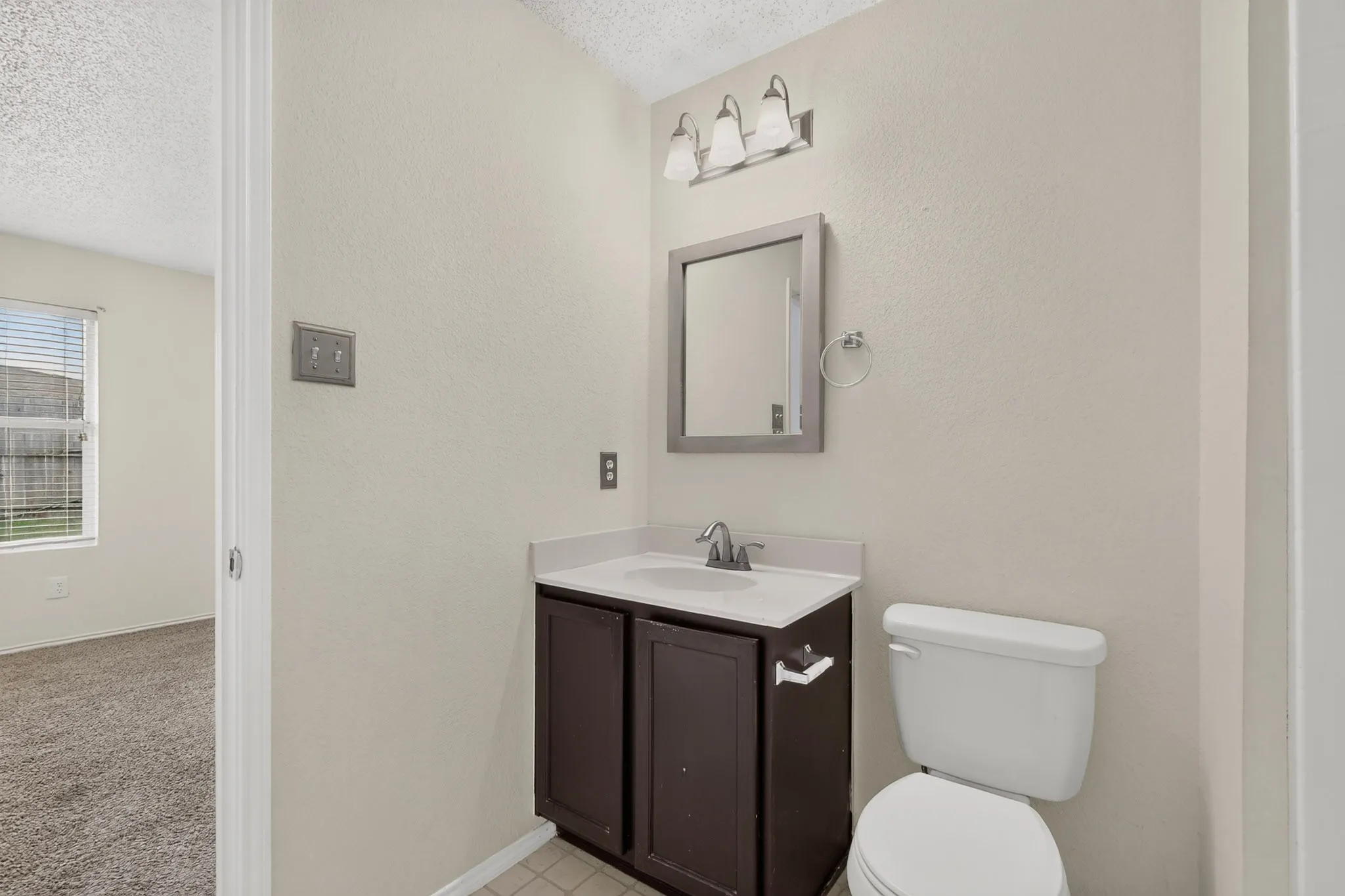 Vanity with dark cabinetry and white countertop, integrated sink, wall-mounted mirror, three-light fixture, and porcelain toilet