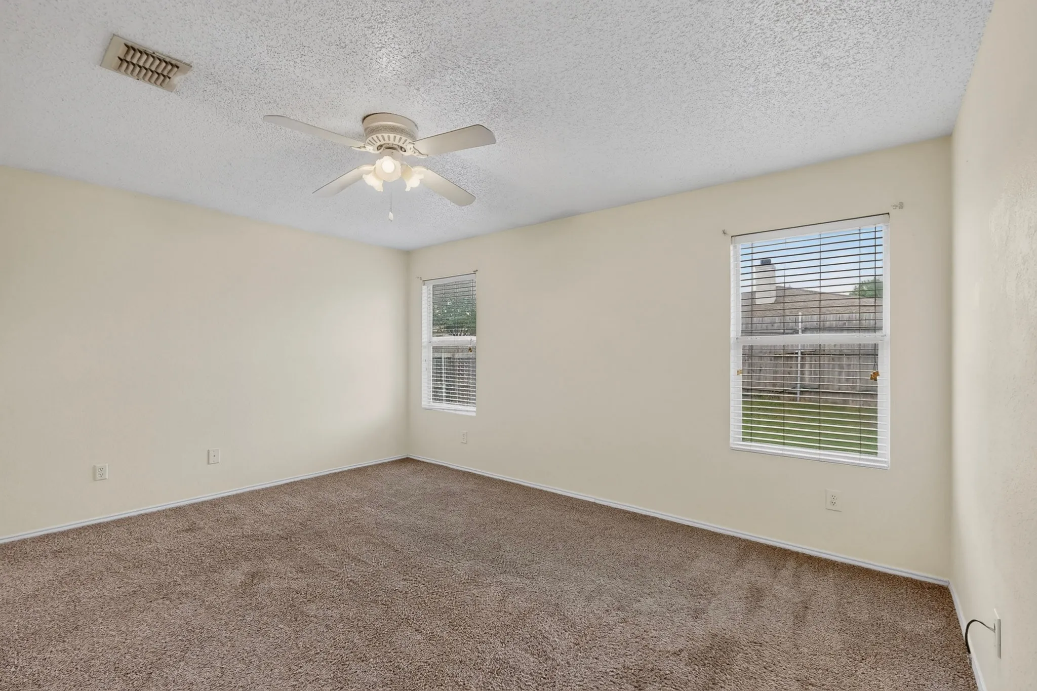 Neutral-toned interior space featuring carpeted flooring, two windows with horizontal blinds, and a ceiling-mounted light fixture with a fan