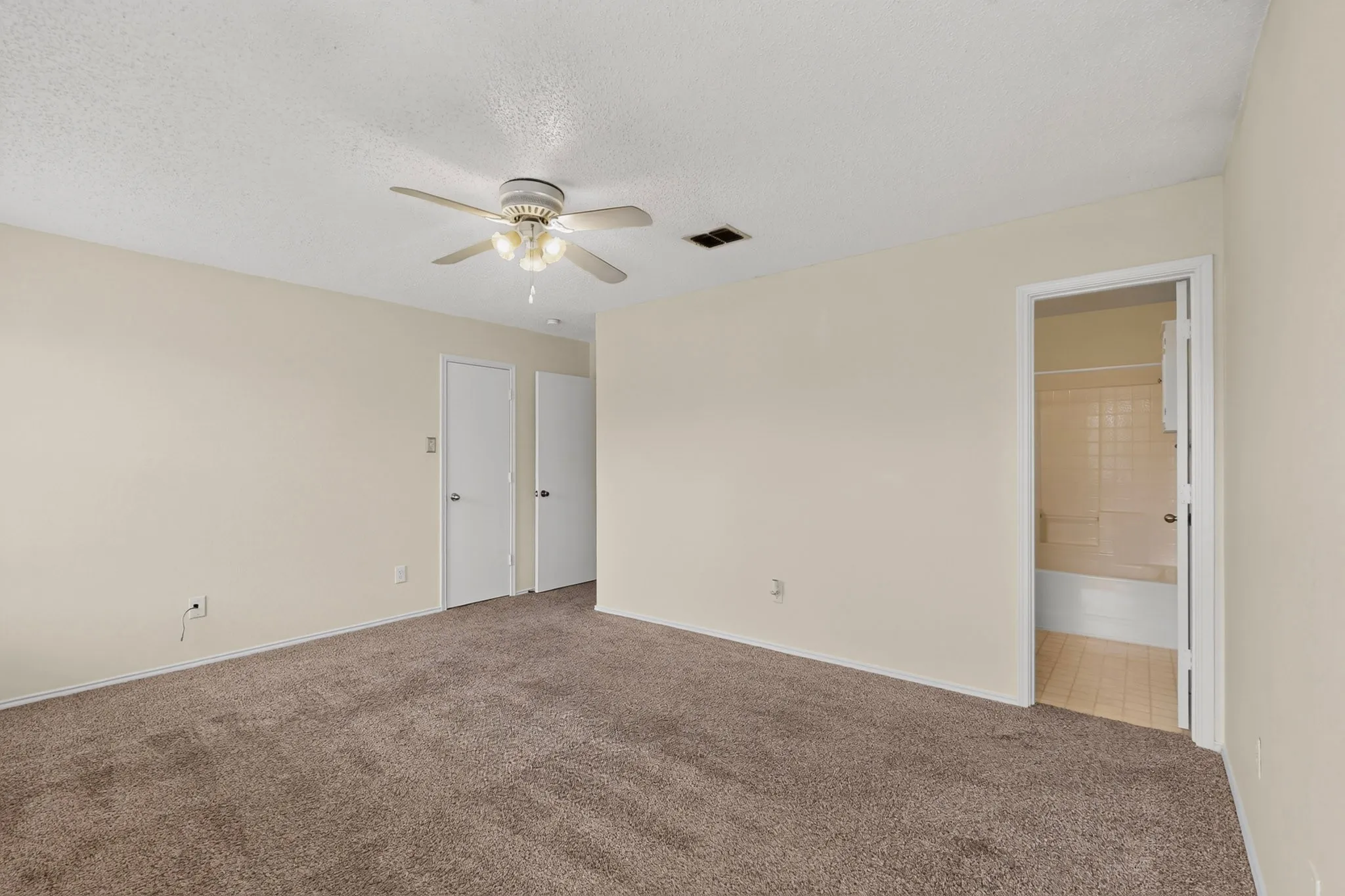 Neutral-toned interior space featuring carpeting, a ceiling fan with integrated lighting, two interior doors, and a doorway opening into a tiled bathroom