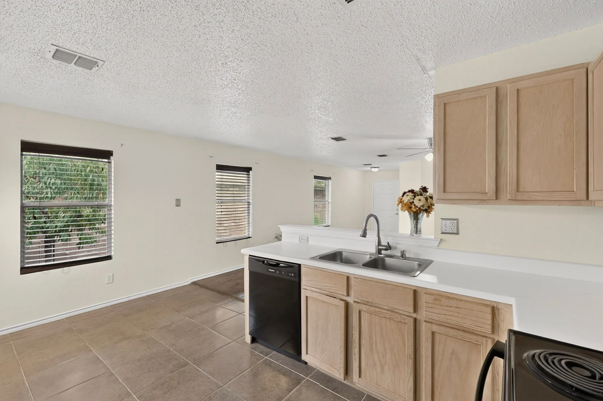 Kitchen featuring light wood-finish cabinetry, solid surface countertops, dual-basin stainless steel sink, black built-in dishwasher, and ceramic tile flooring