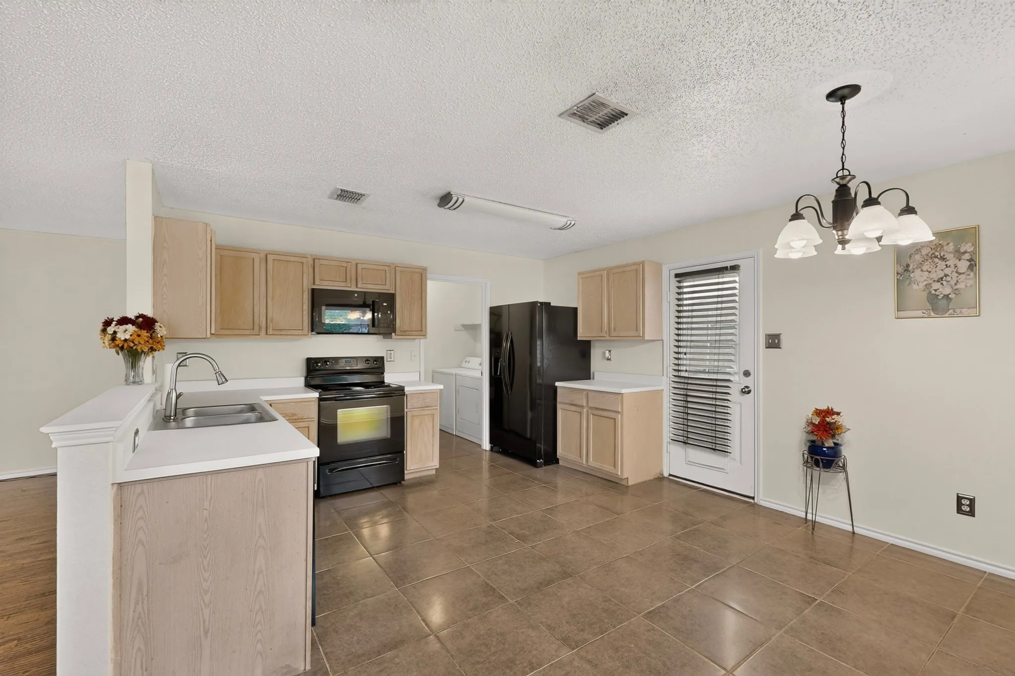 Kitchen featuring light-finish cabinetry, neutral-toned tile flooring, integrated black appliances, and a traditional multi-light chandelier
