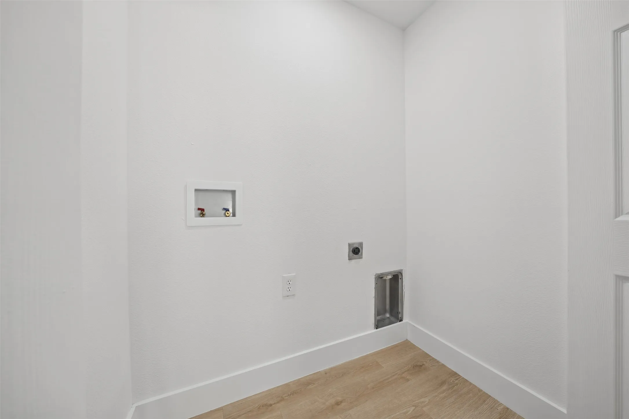 Utility area featuring wood-finish flooring, white textured walls, recessed water hookups, and integrated electrical and ventilation outlets