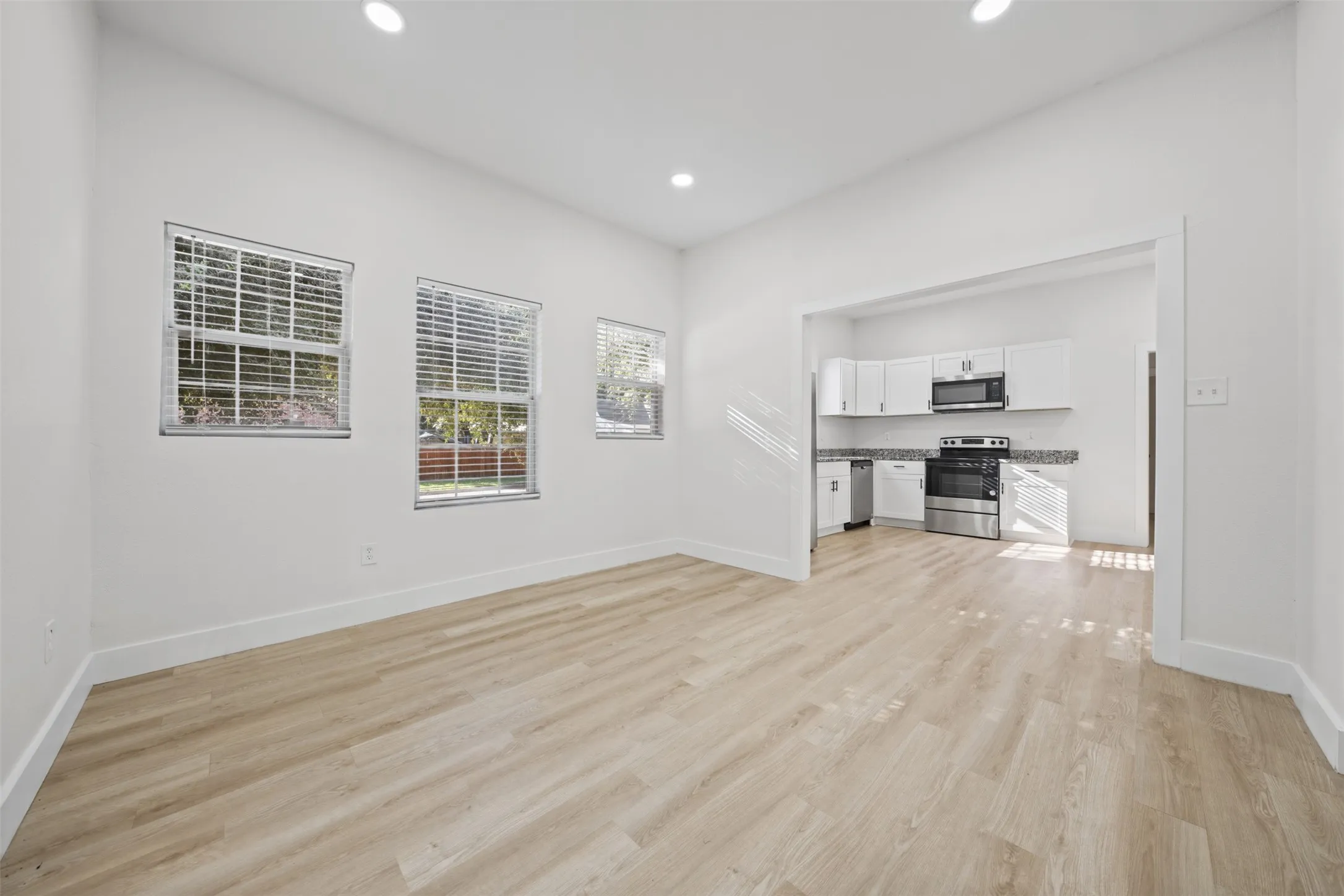 Open-concept interior featuring wood-finish flooring, recessed lighting, three multi-pane windows, and a kitchen equipped with white cabinetry and stainless steel appliances