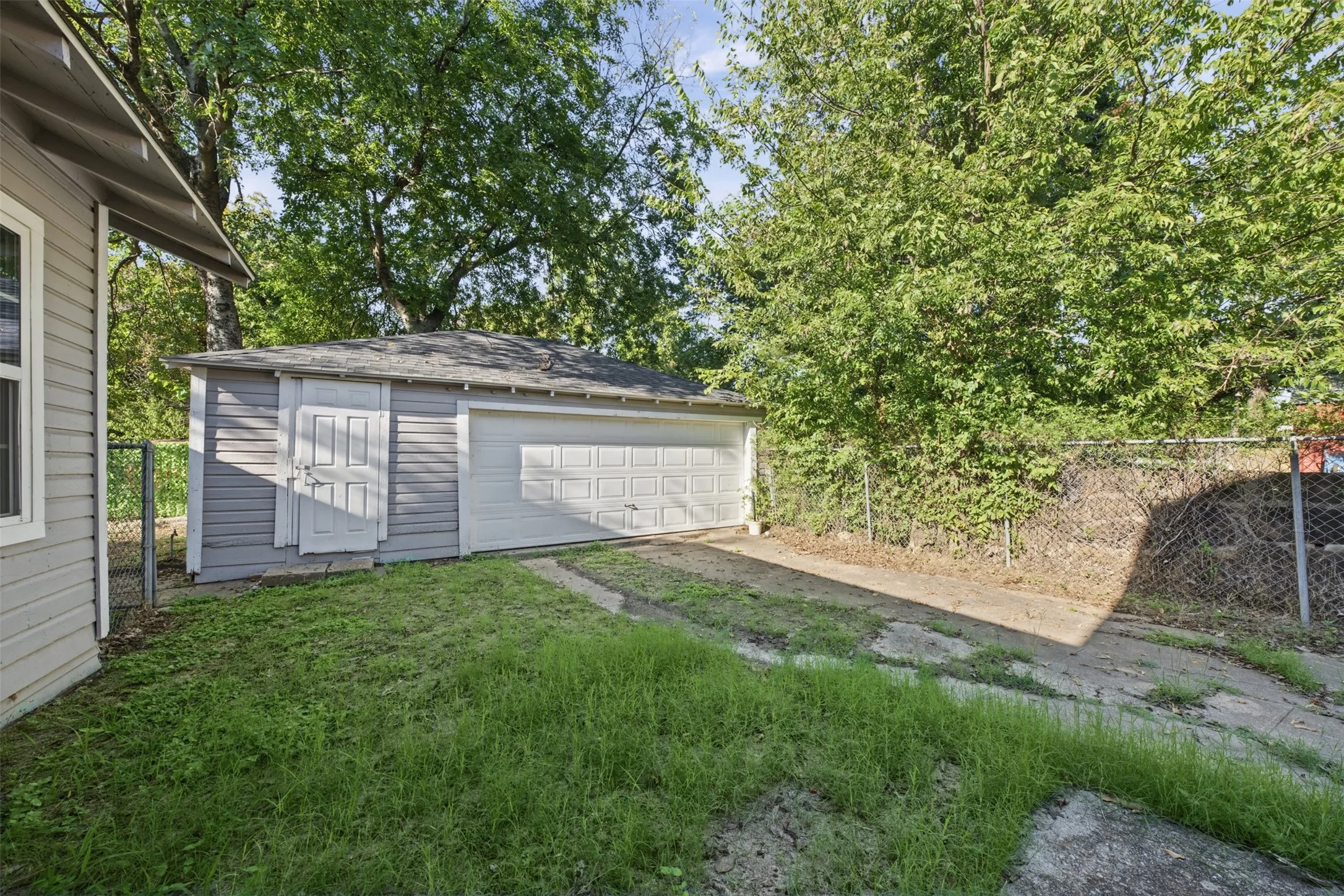 Detached garage with horizontal siding, a roll-up sectional door, and an individual side entry door, situated on a lot with concrete hardscaping and perimeter chain-link fencing