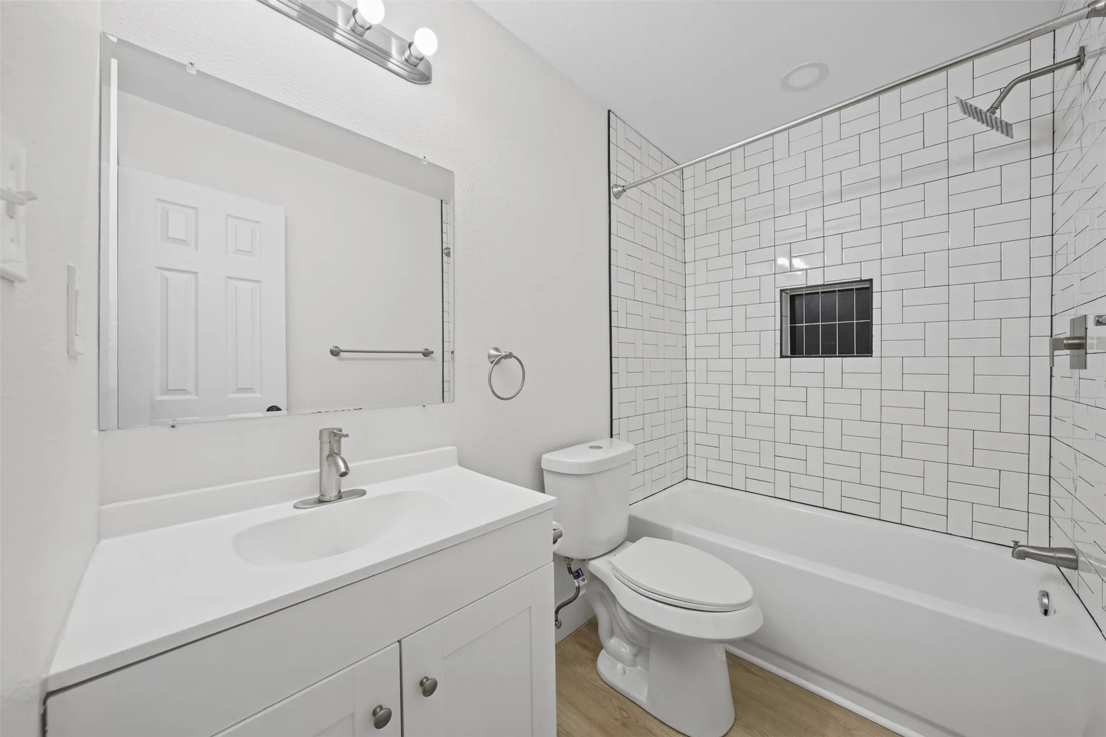 Bathroom featuring a white vanity with an integrated sink, polished chrome faucet, rectangular framed mirror, wood-finish flooring, and a bathtub enclosure with white subway tile and a built-in recessed wall niche