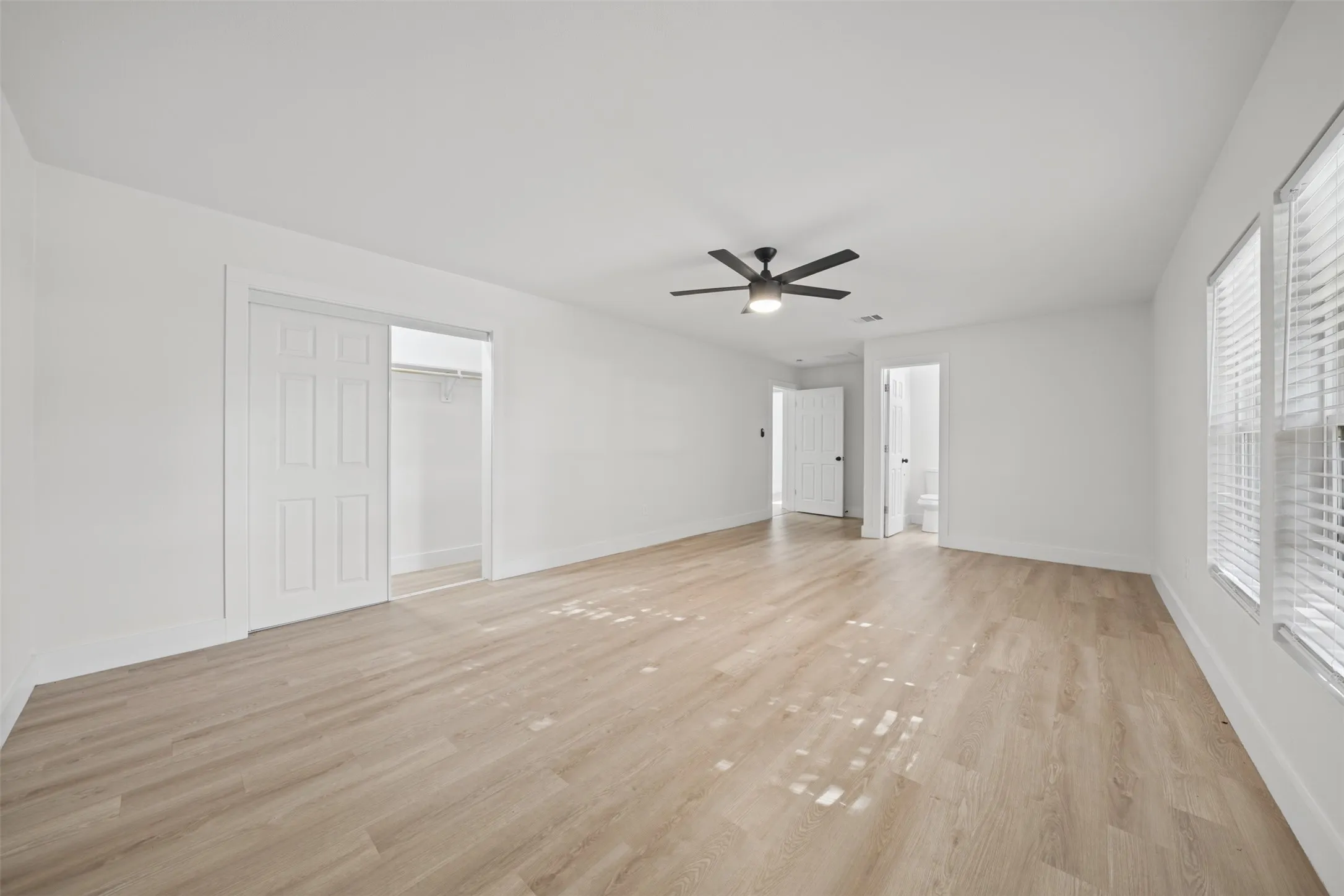 Interior room featuring wood-finish flooring, a matte black ceiling fan with integrated lighting, white walls, and a walk-in closet with built-in shelving