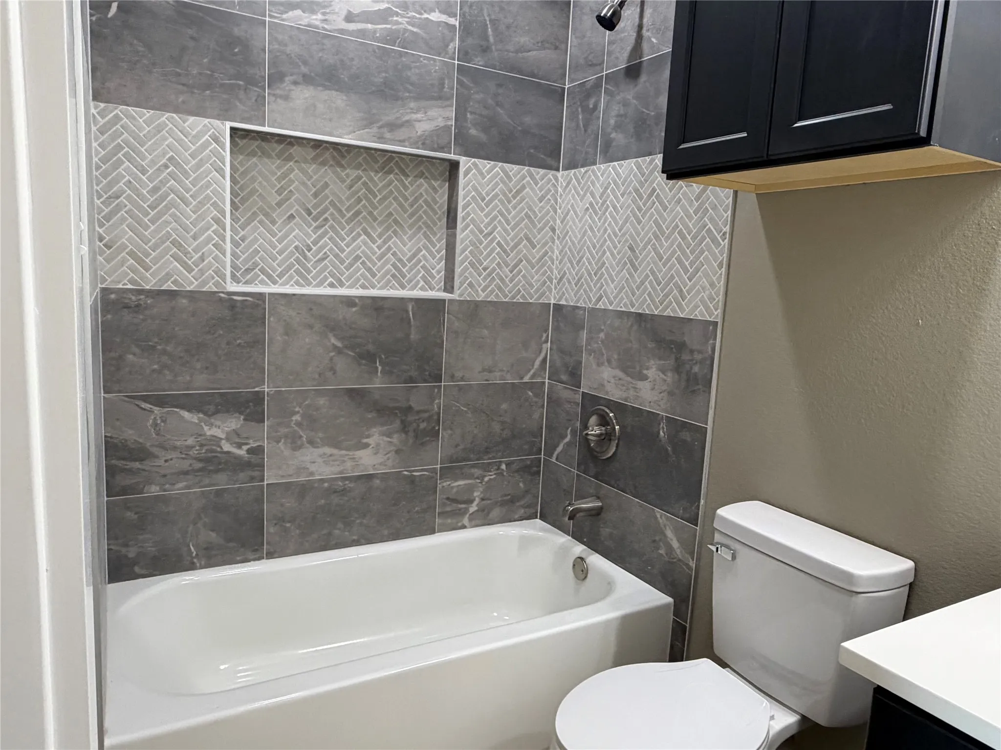 2nd Bed bath Bathtub featuring large-format grey tile surround, herringbone tile inlay, integrated shelving niche, polished chrome fixtures, and wall-mounted black cabinetry