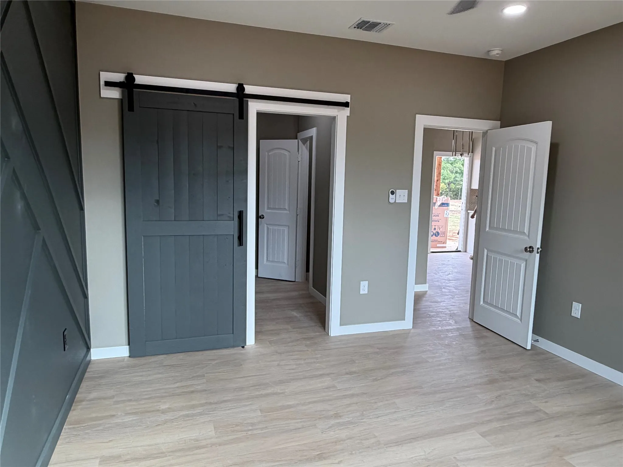 Interior space featuring light wood-finish flooring, a charcoal gray barn door with black hardware, white paneled interior doors, and a charcoal gray accent wall with geometric wood trim