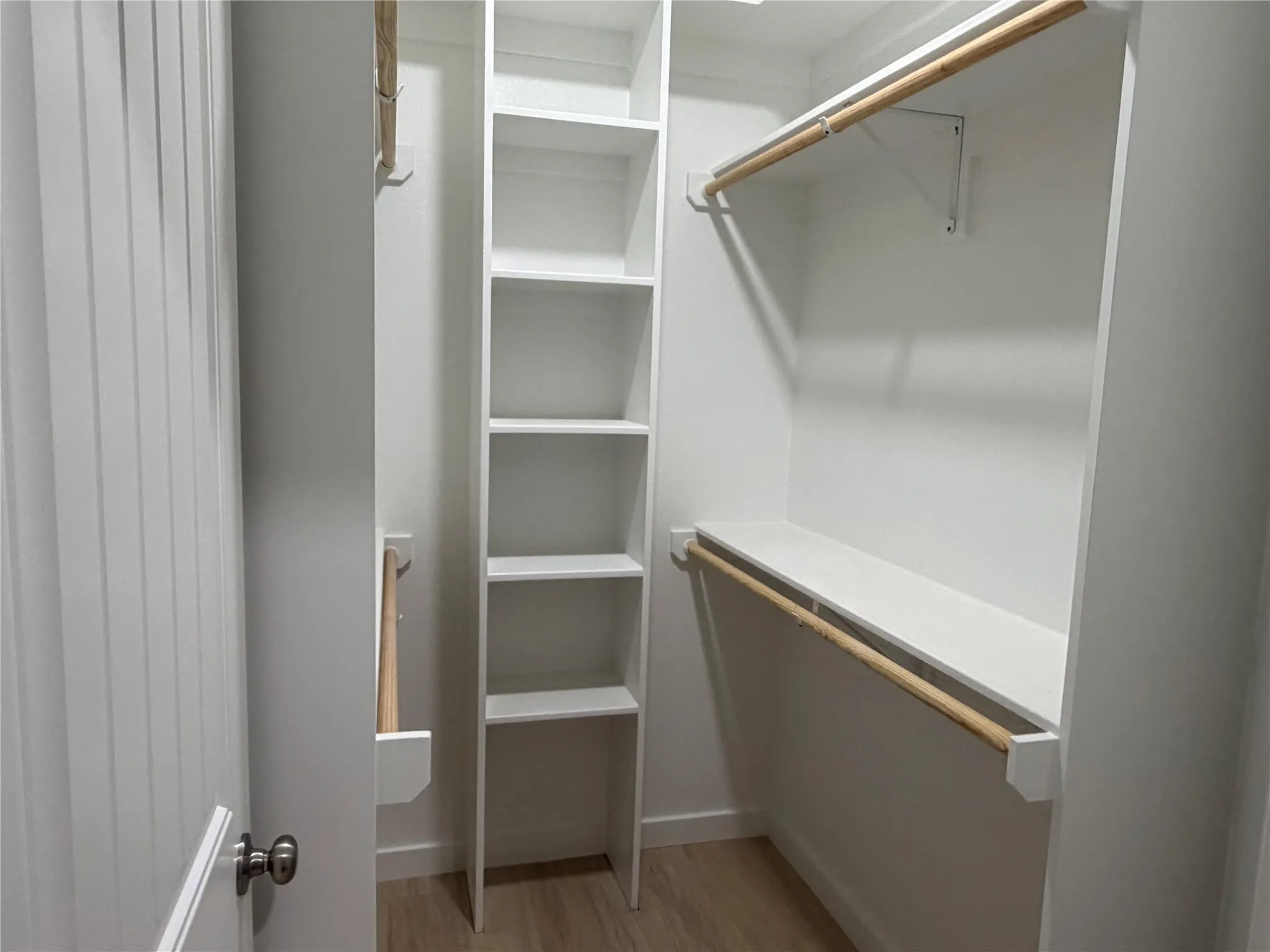 Primary Walk-in closet featuring white built-in shelving, wooden garment rods, and light wood-finish flooring