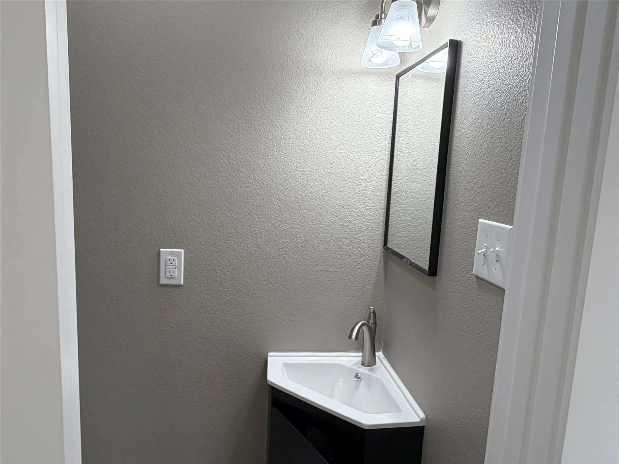 1/2 Bath Corner vanity featuring a triangular basin, metallic faucet, wall-mounted rectangular mirror, and multi-bulb light fixture