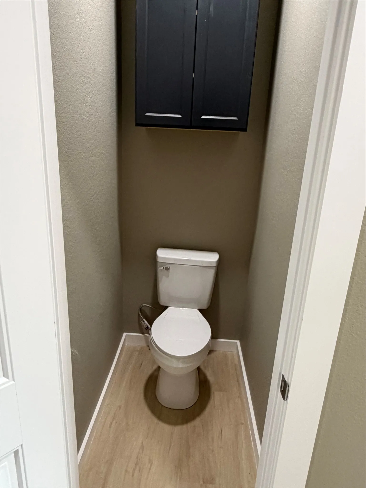 Wall-mounted dark-toned cabinetry, standard white porcelain toilet, and light-toned wood-finish flooring