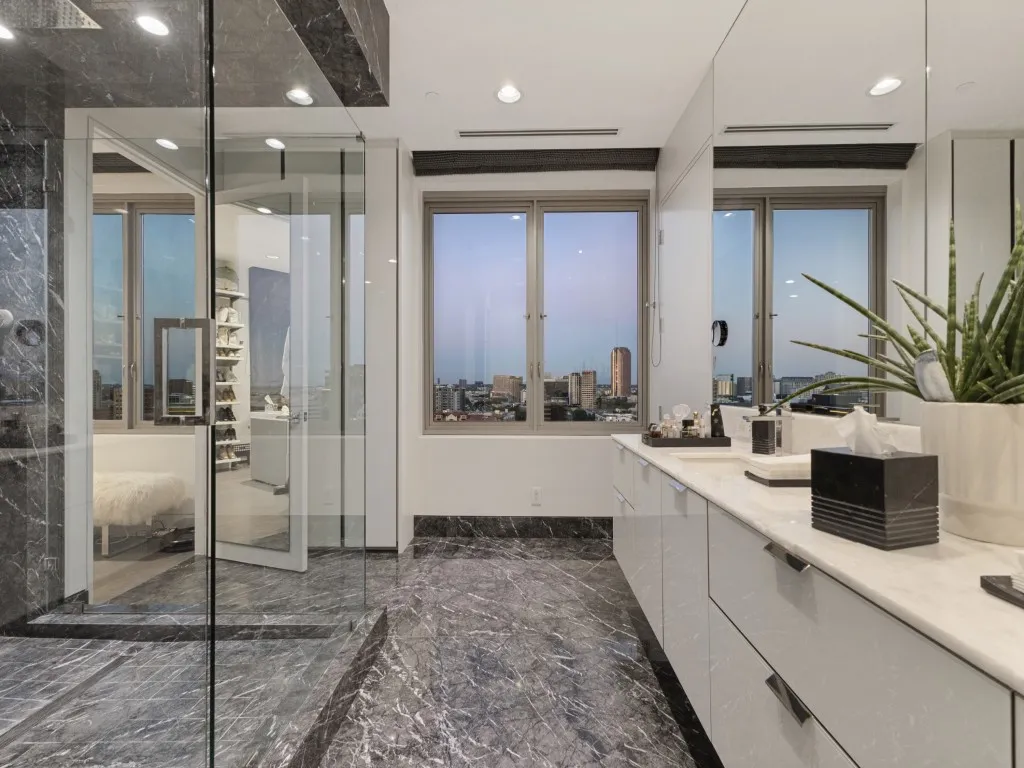 Shower enclosure with glass paneling, grey marble-finish flooring, dual-sink vanity with white countertop, and expansive windows