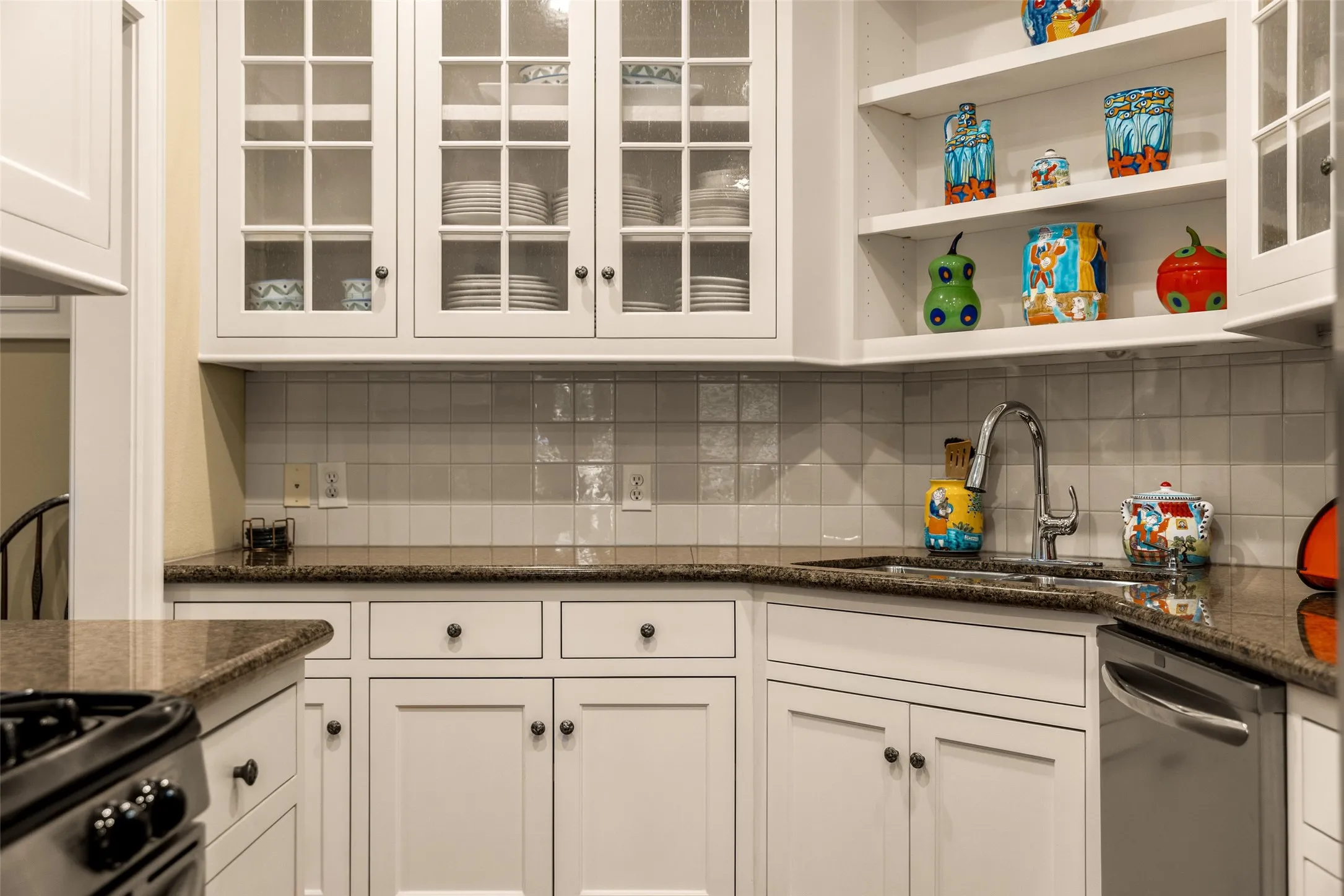 White cabinetry featuring glass-panel upper doors, open shelving, stone countertops, square-tile backsplash, and stainless steel sink hardware