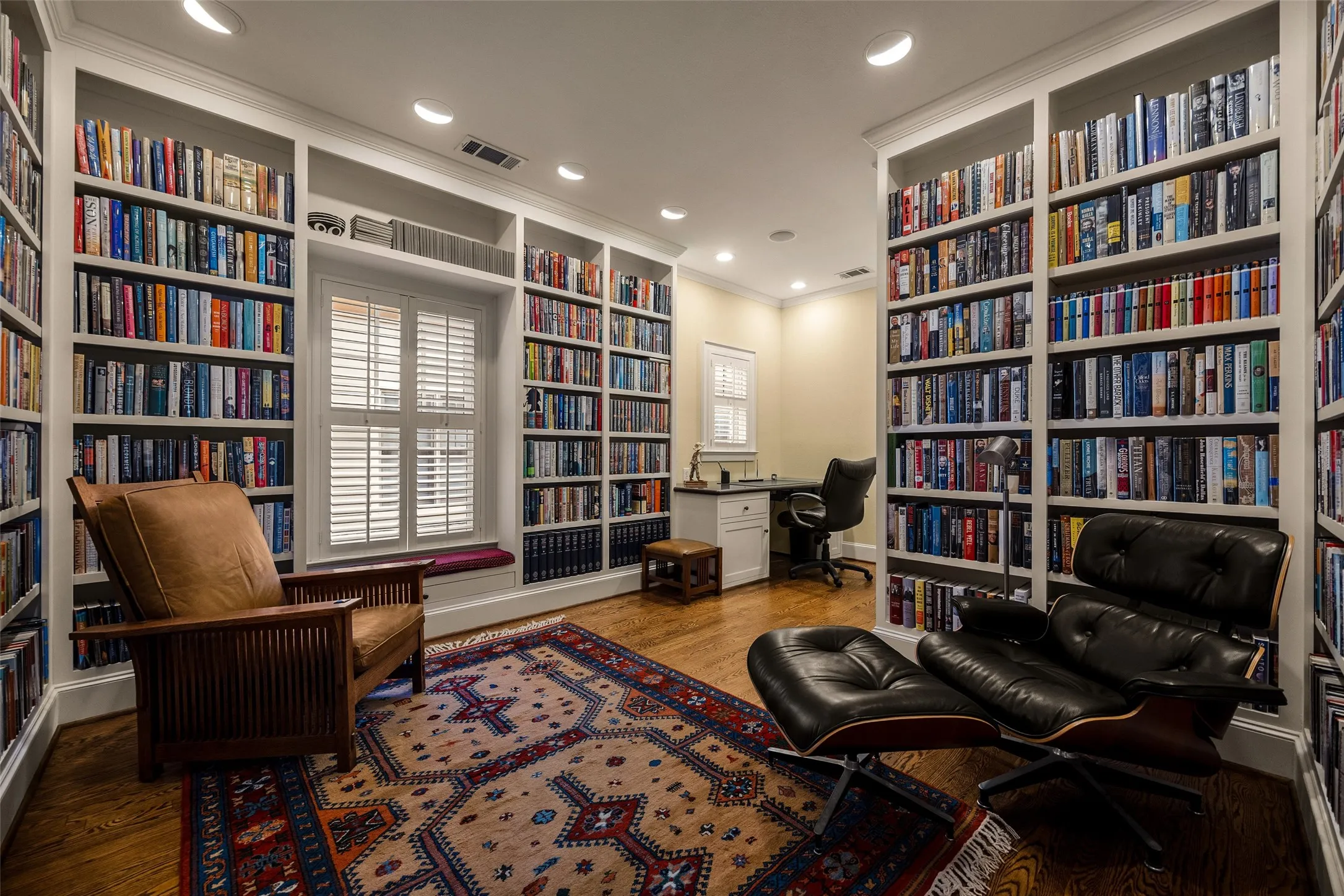 Floor-to-ceiling custom built-in shelving, hardwood flooring, recessed lighting, integrated window seat, and built-in desk