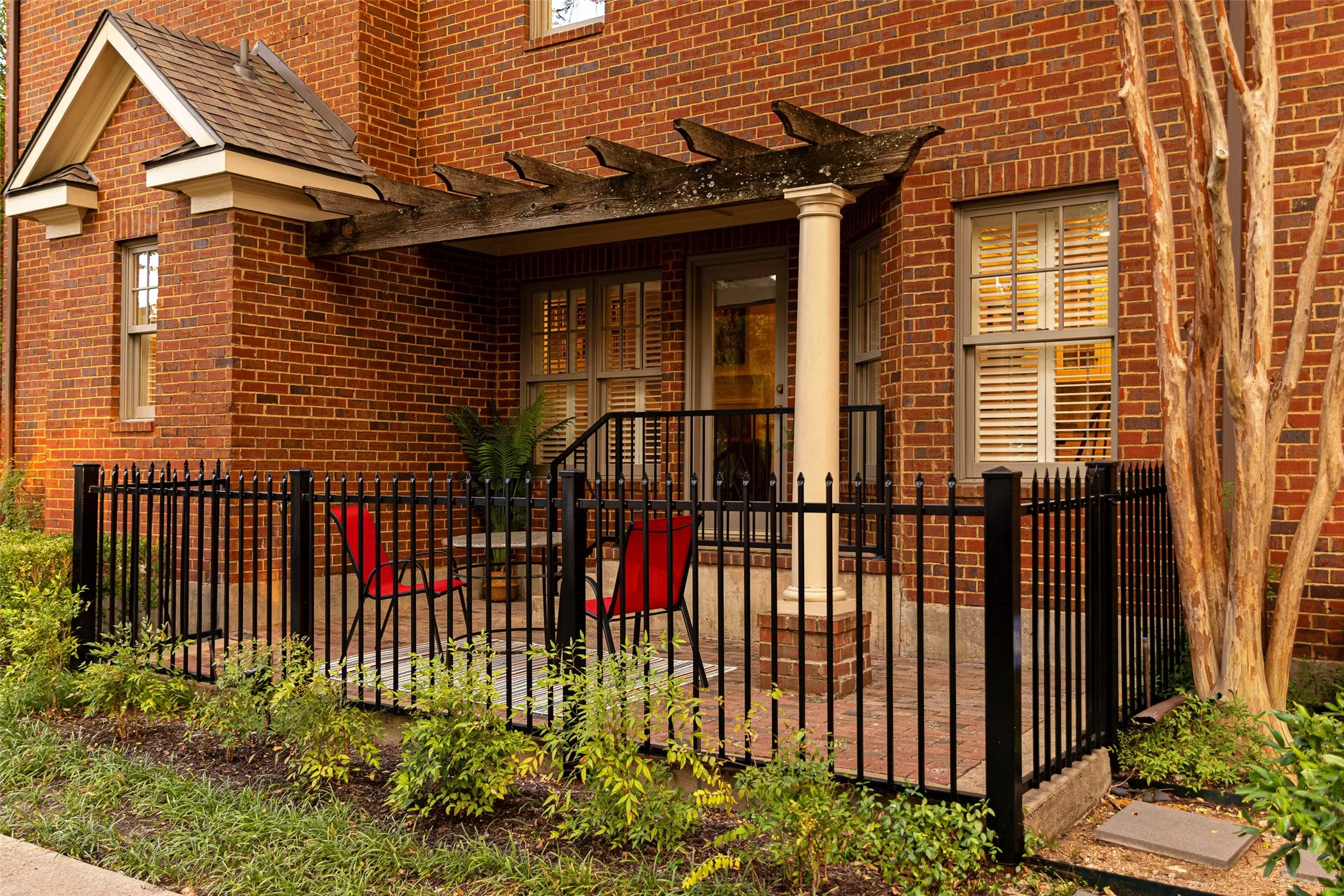 Red brick exterior featuring a covered patio, wooden pergola, decorative column support, and black metal fencing