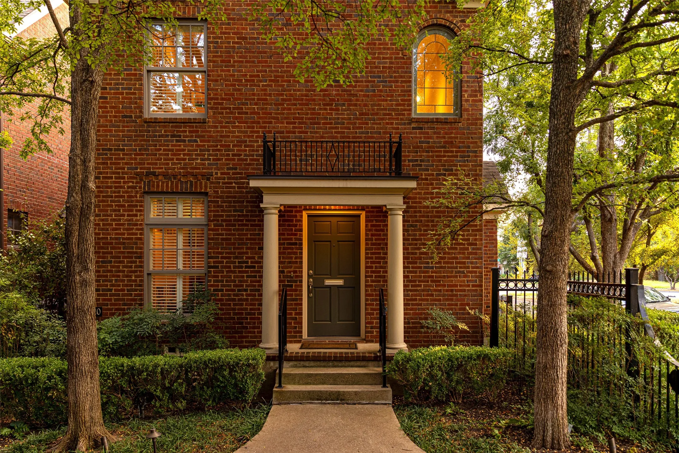 Brick exterior featuring a covered entryway with structural columns, a wrought iron balcony, an arched window, and a paved walkway
