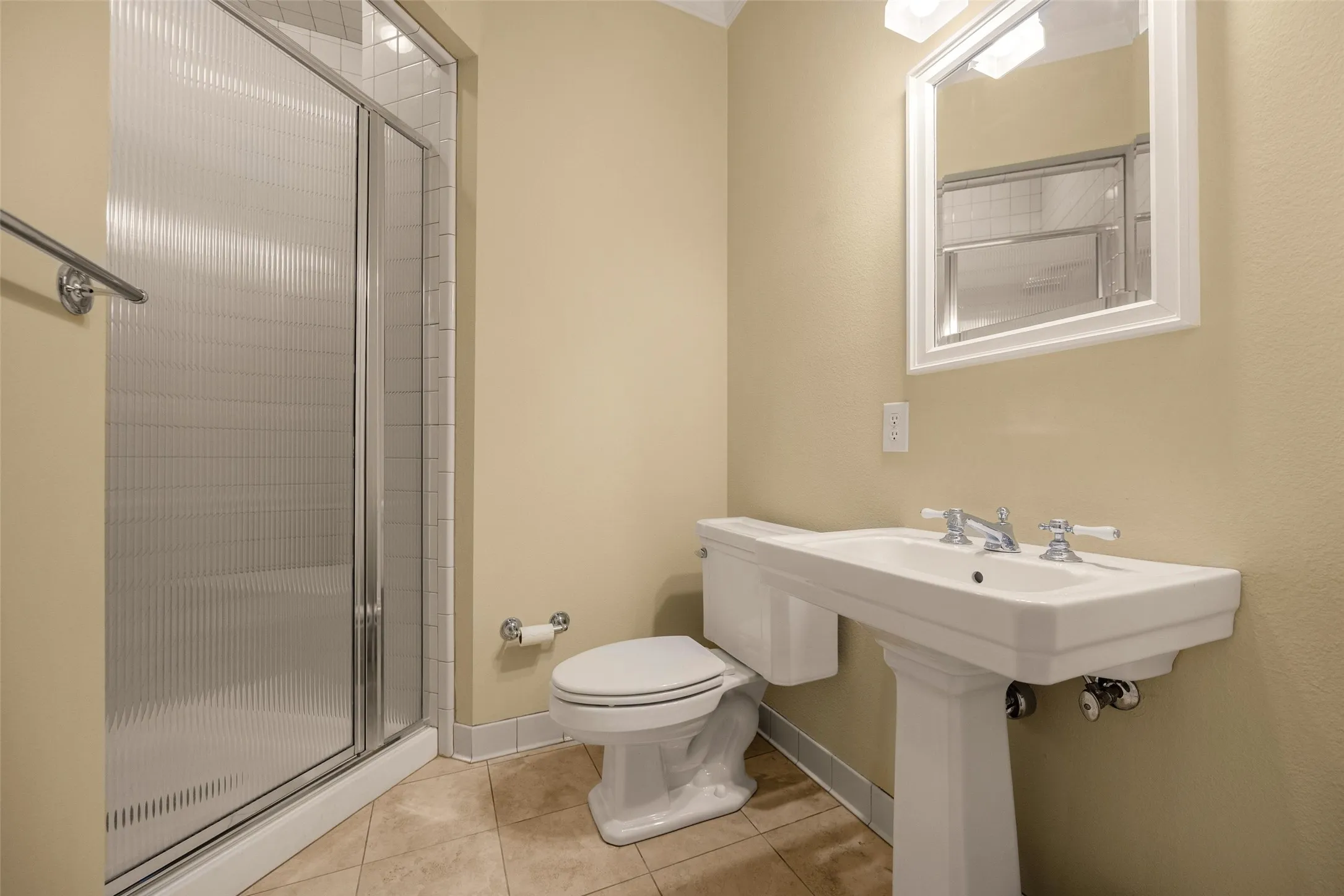 Shower with semi-frameless fluted glass doors and tiled surround, pedestal sink with cross-handle faucet, white framed wall mirror, and neutral-tone tile flooring
