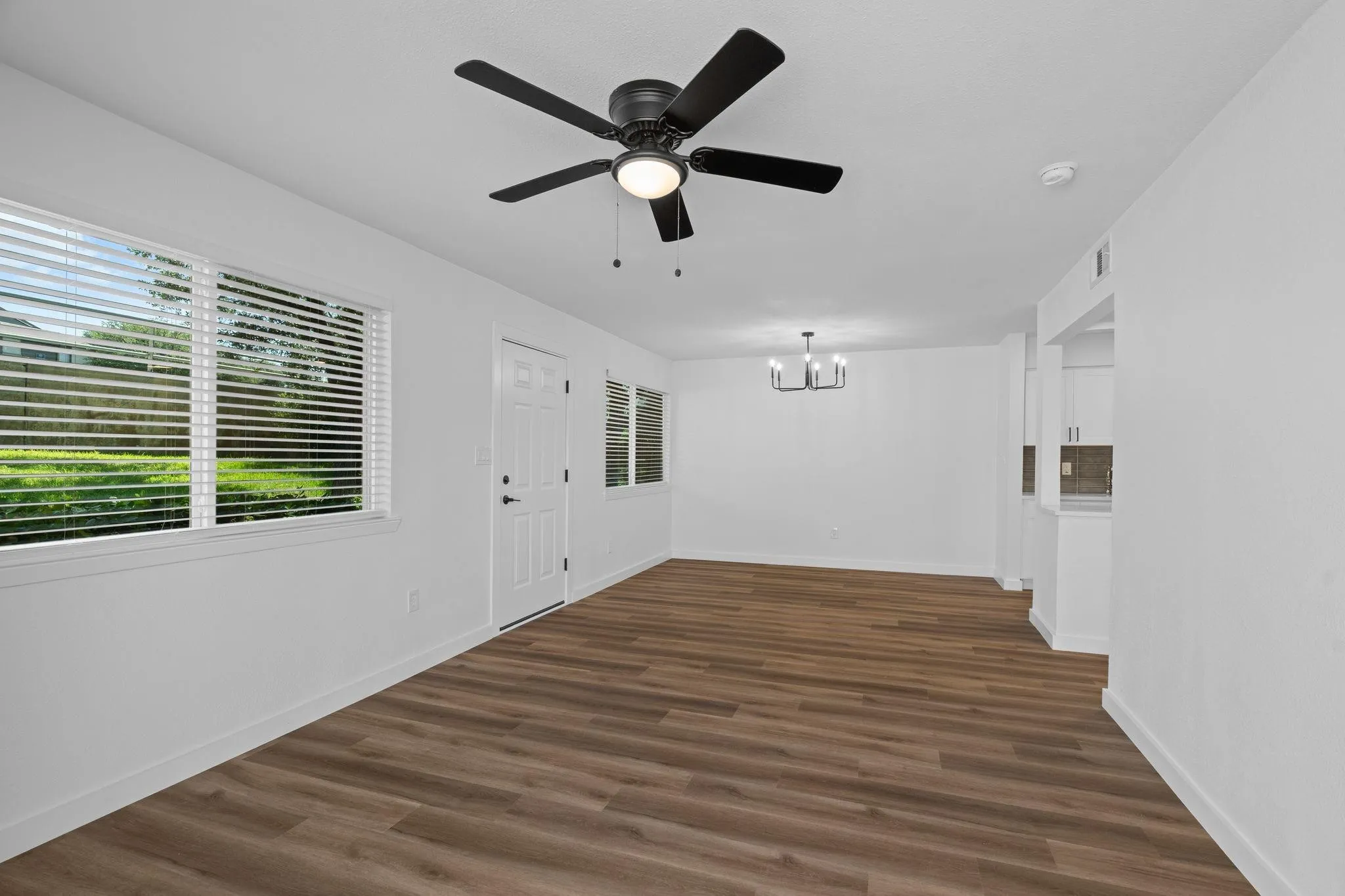 Wood-finish flooring, matte black ceiling fan with integrated lighting, multipane windows with horizontal blinds, and a modern six-light chandelier