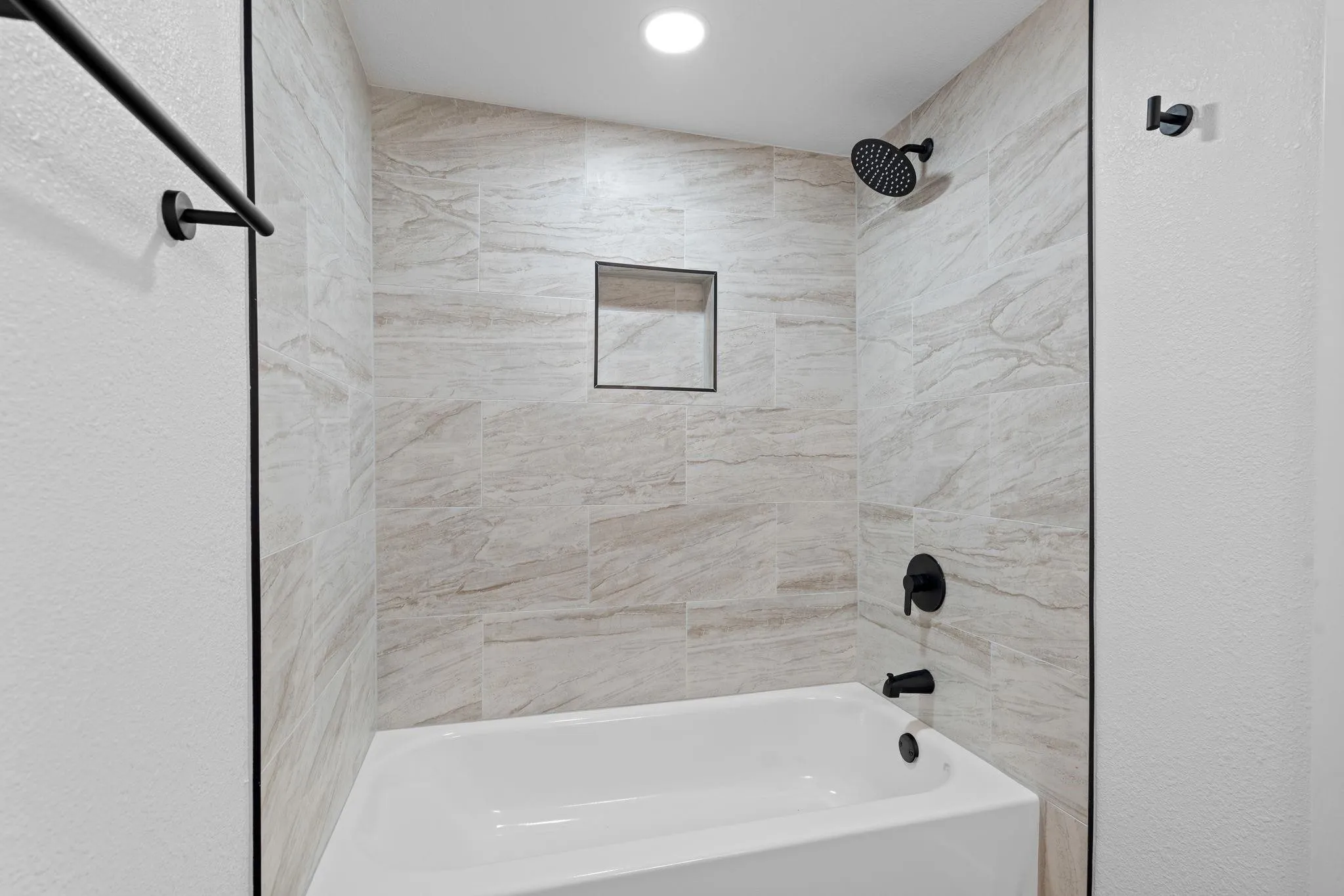 Bathtub surround featuring large-format neutral-toned tile, recessed wall niche, matte black fixtures, and a shower head